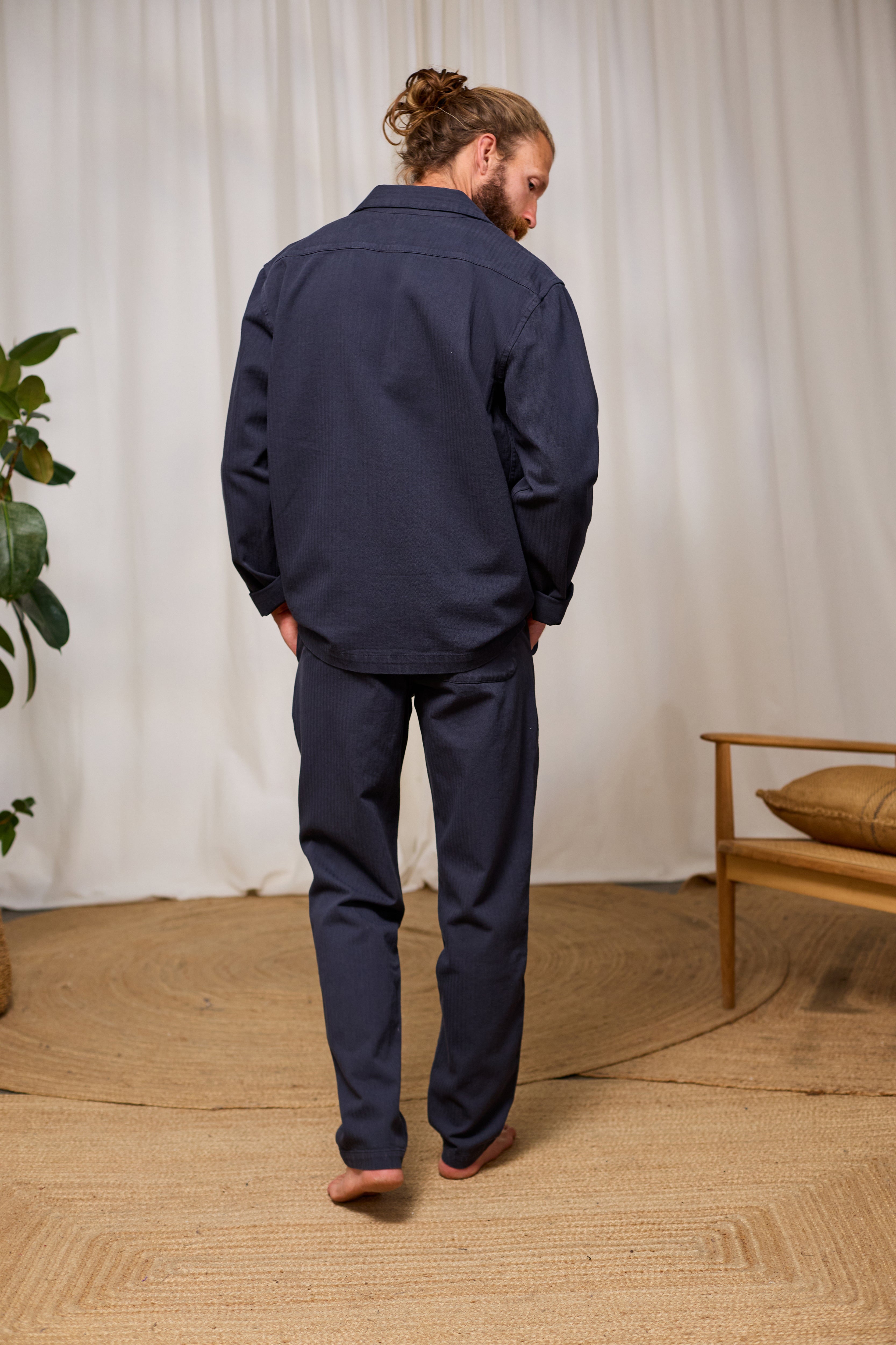 Man wearing a navy blue outfit walking in a room with a plant and wooden furniture.