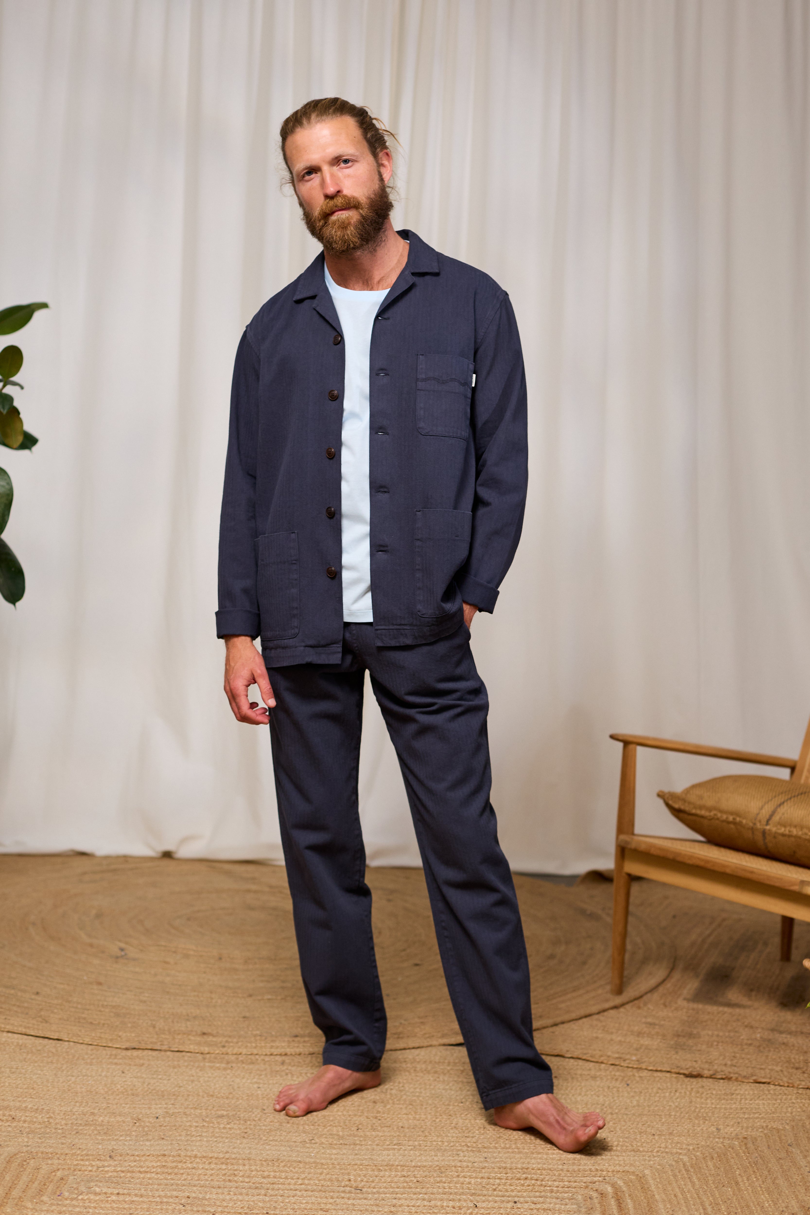 Man wearing a navy blue shirt and pants standing in a room with a wooden chair and plant.