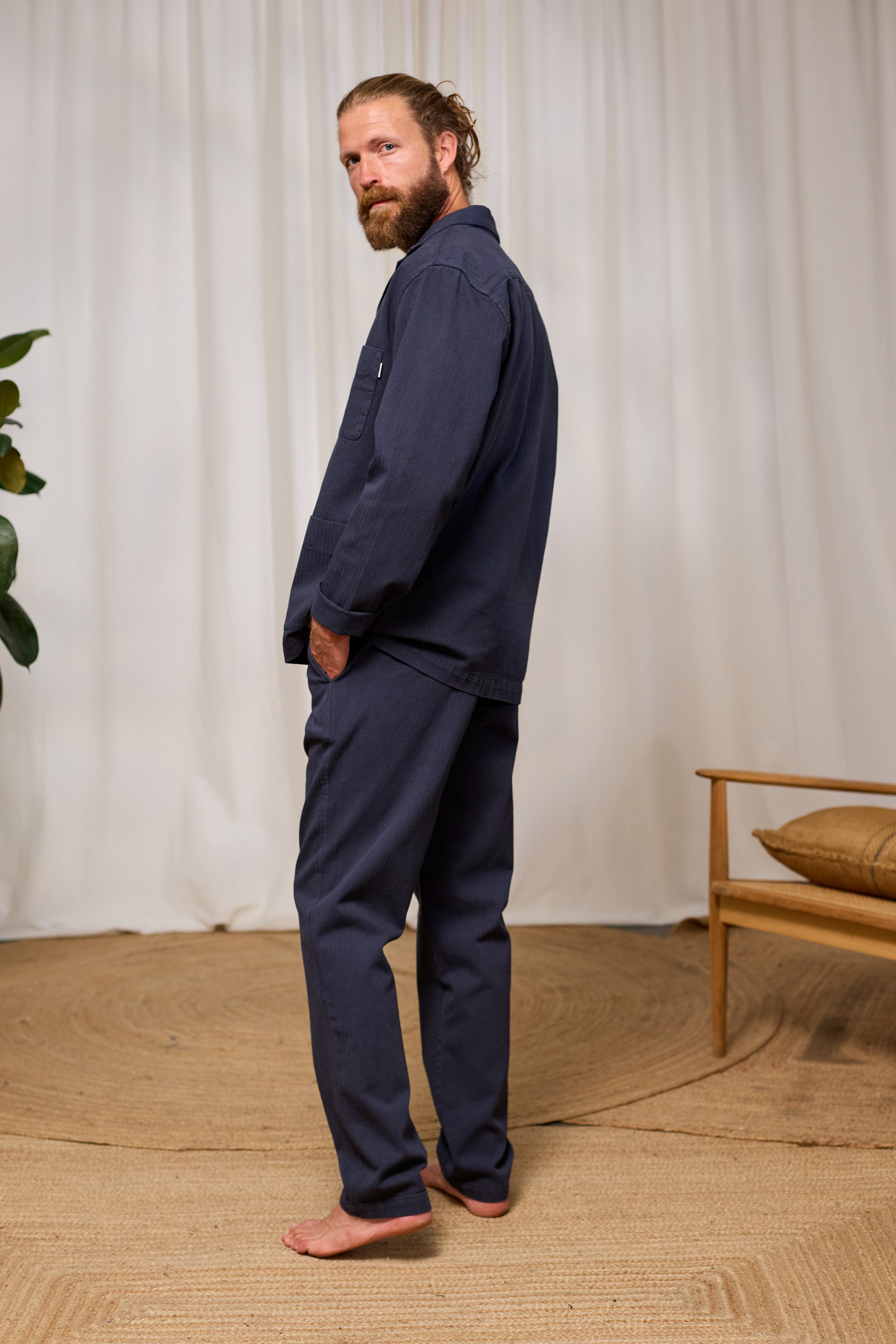 Man wearing a navy blue organic cotton outfit standing in a room with a white curtain and wooden furniture.