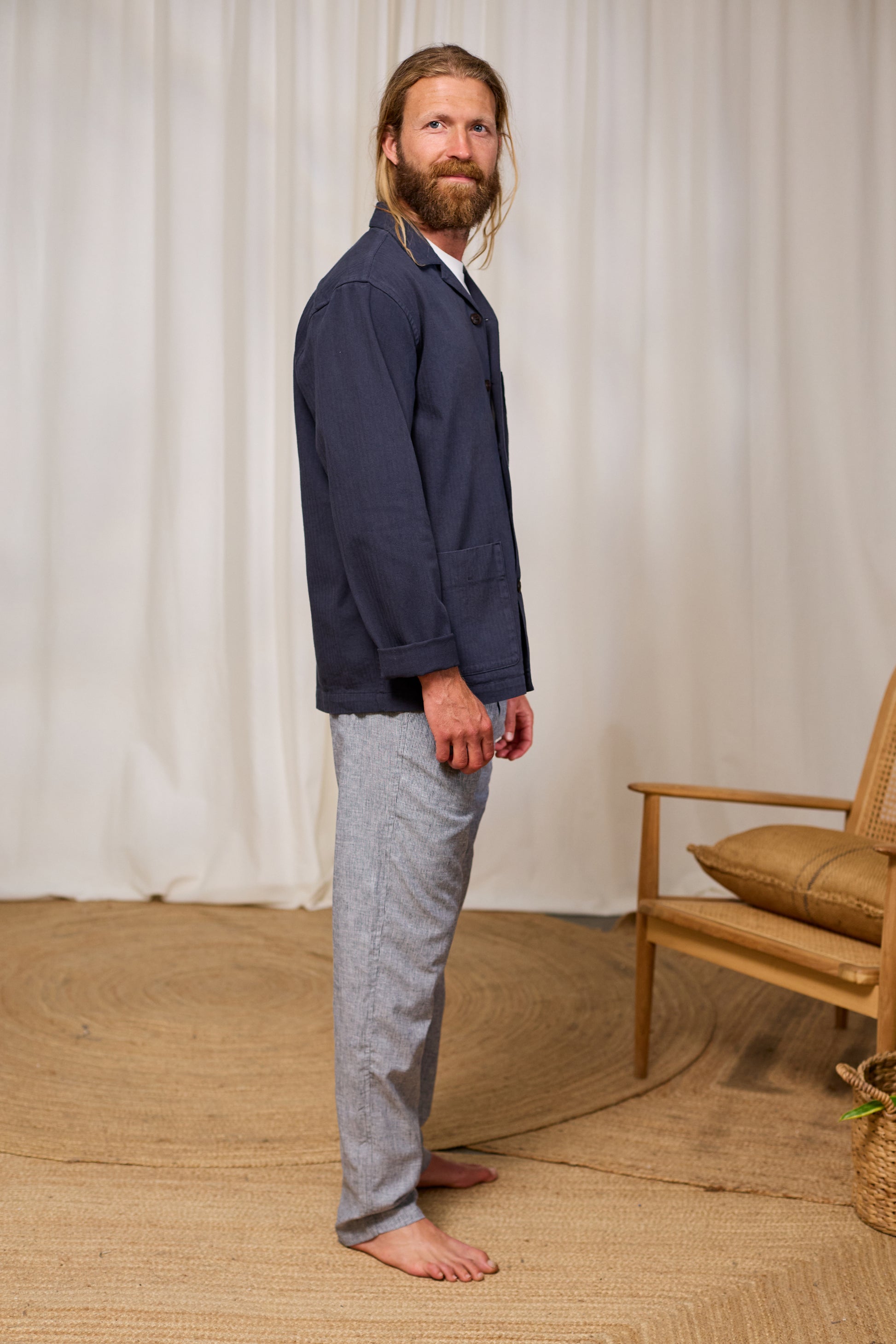 Man wearing a navy shirt and gray pants standing in a room with a wooden chair and woven rug.