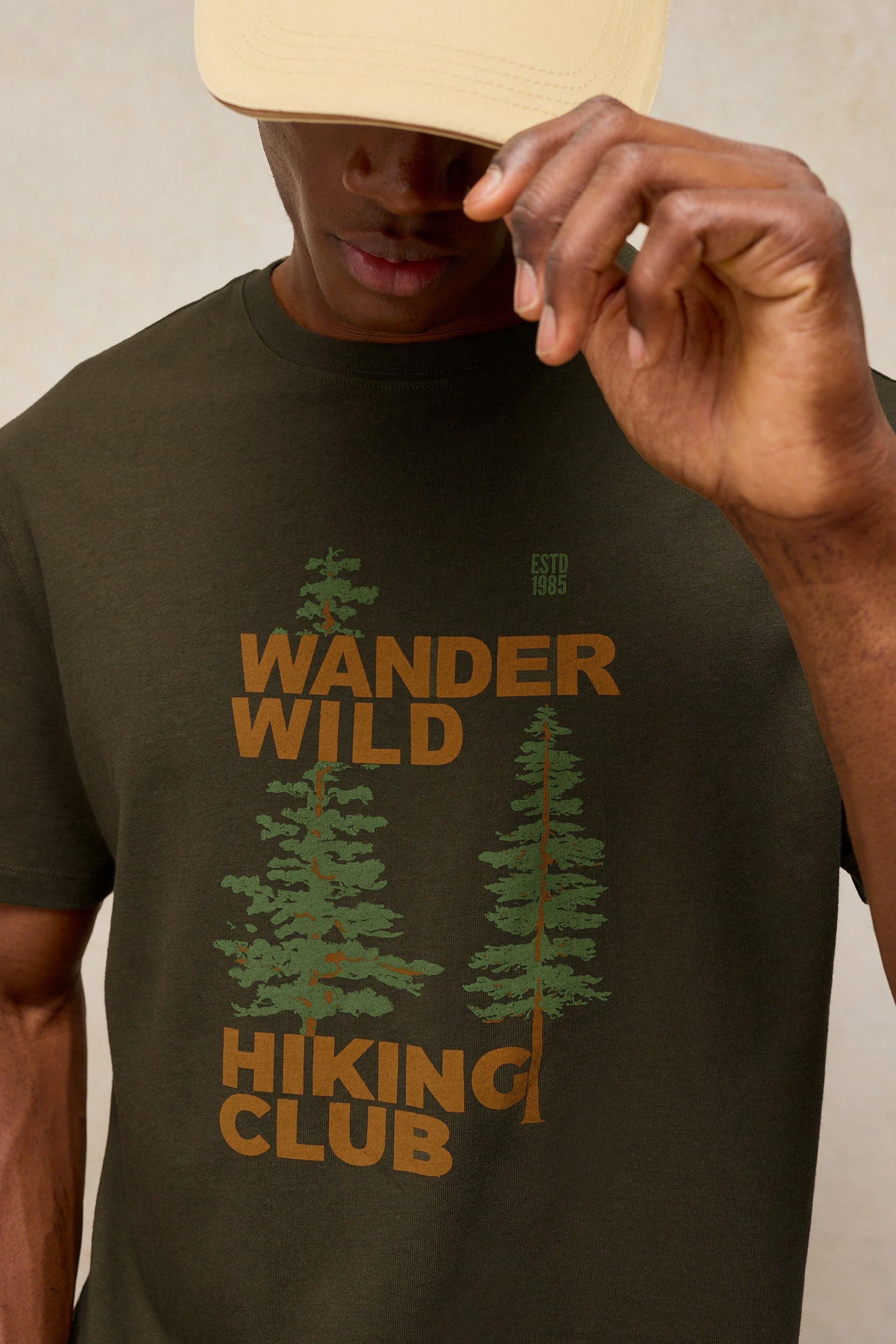 A person wearing a dark olive green T-shirt with Wander Wild Hiking Club graphic text and pine tree illustrations