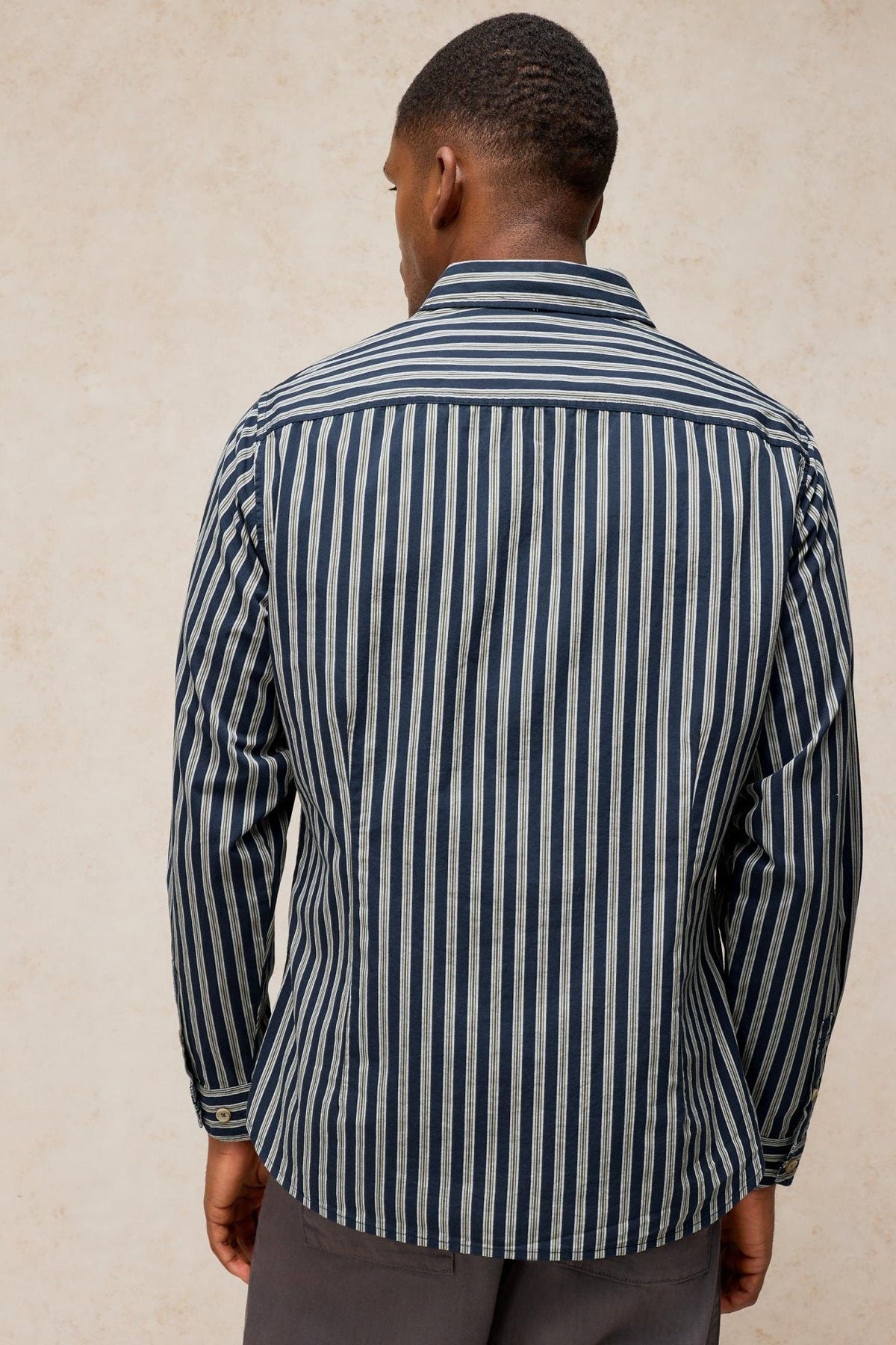 Rear view of a man wearing a long sleeve White Stuff navy and white striped cotton shirt against a plain background.