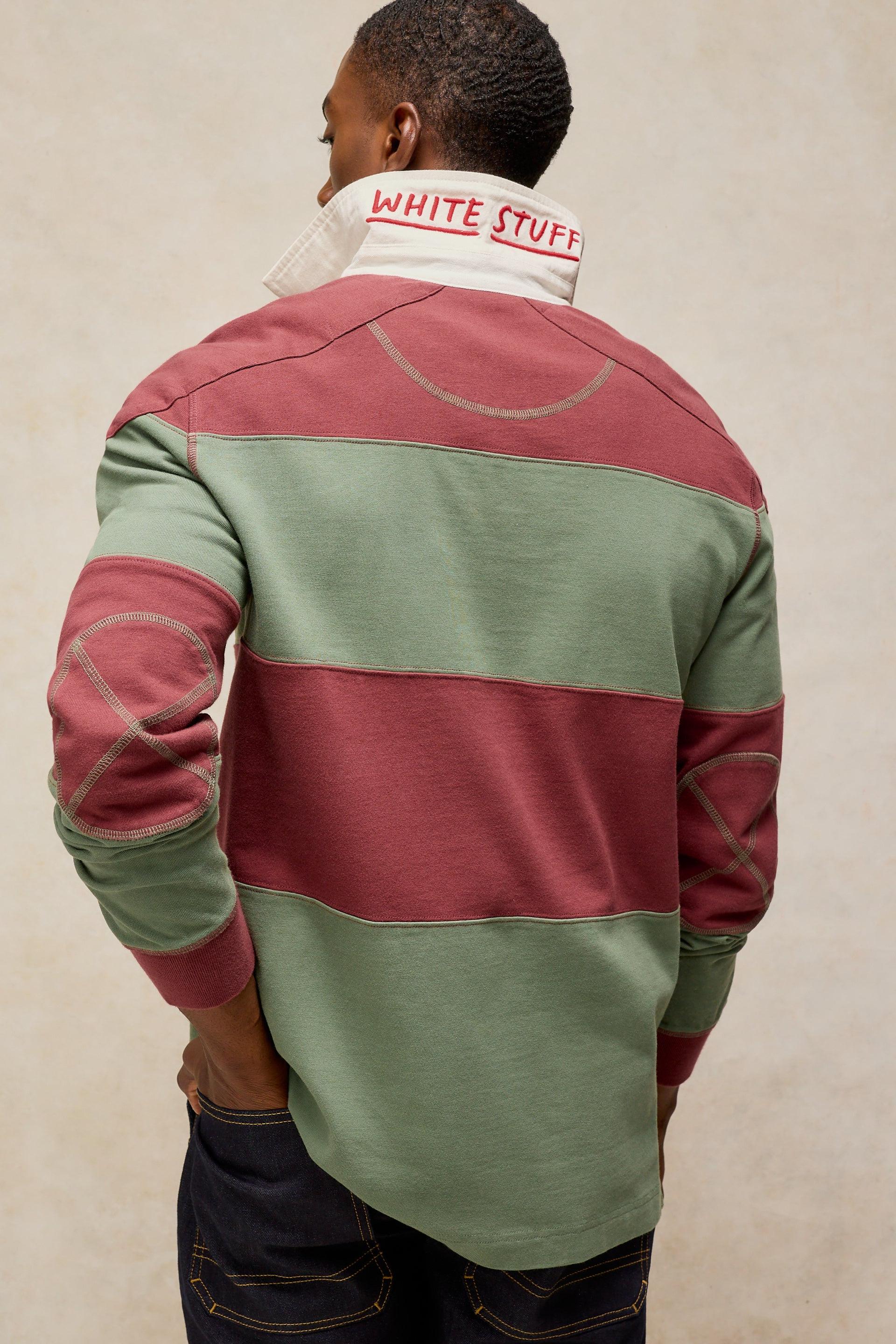 A man viewed from behind wearing a color-blocked rugby-style sweater with horizontal stripes in sage green and dusty rose, featuring contrast stitching and a white collar with red text reading 'WHITE STUFF'.
