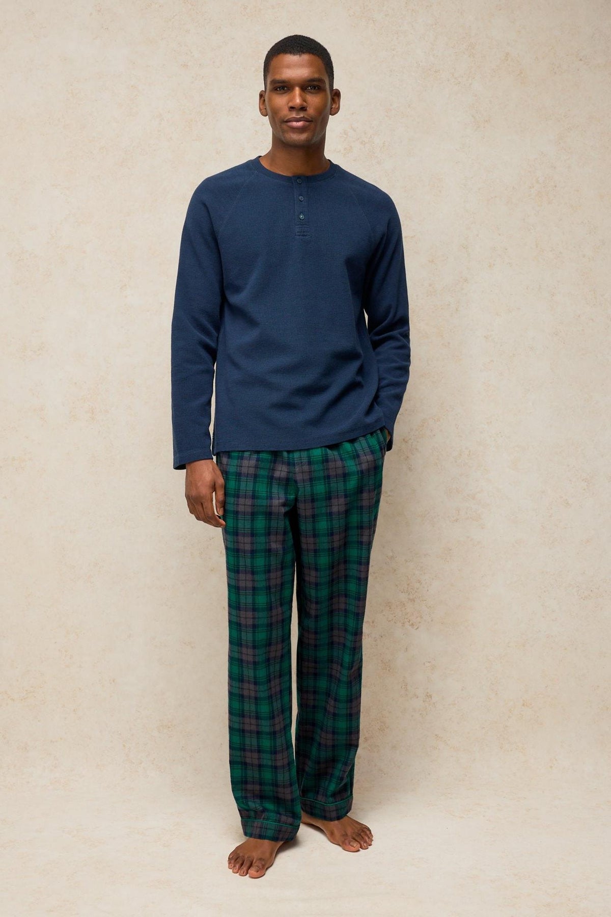A man wearing a navy long-sleeve henley shirt and green plaid White Stuff pyjama trousers standing against a beige wall.