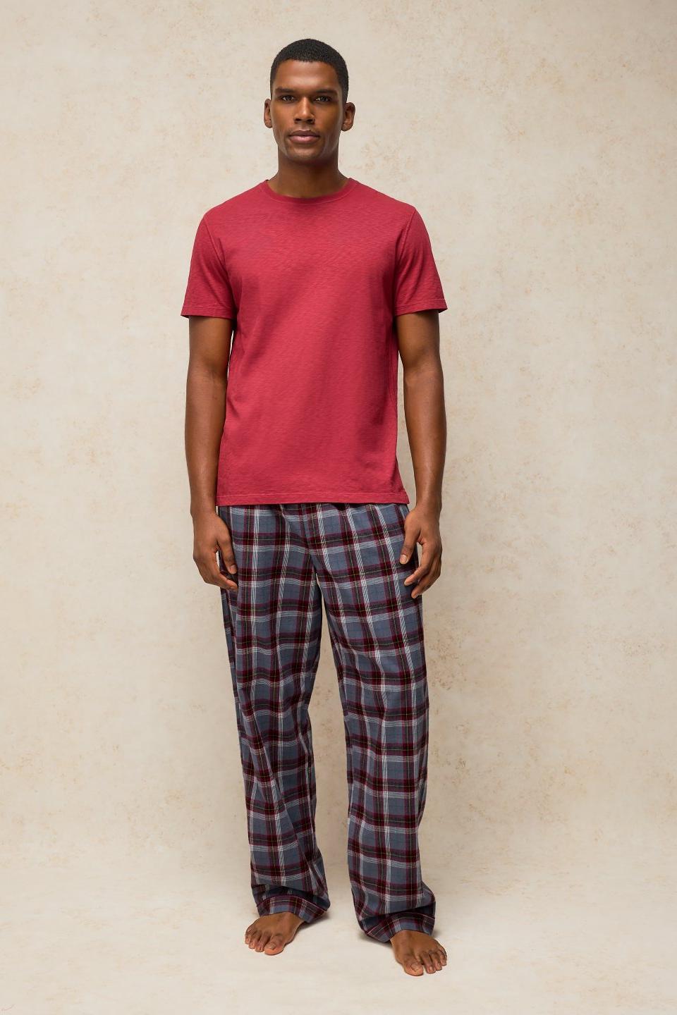 A man wearing a red pyjama top and organic cotton plum check pyjama bottoms against a plain studio background.