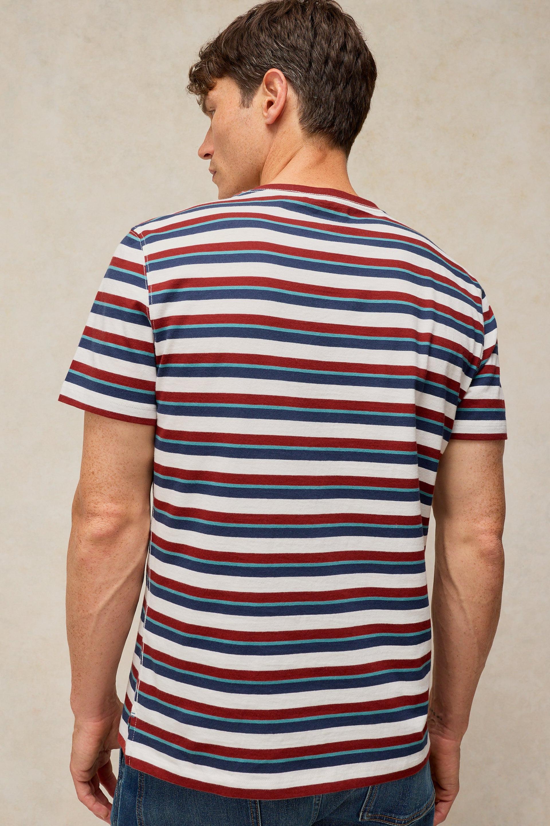 A man viewed from the back wearing a short-sleeve crewneck t-shirt with horizontal red, navy, and white stripes, paired with blue jeans against a textured beige background.