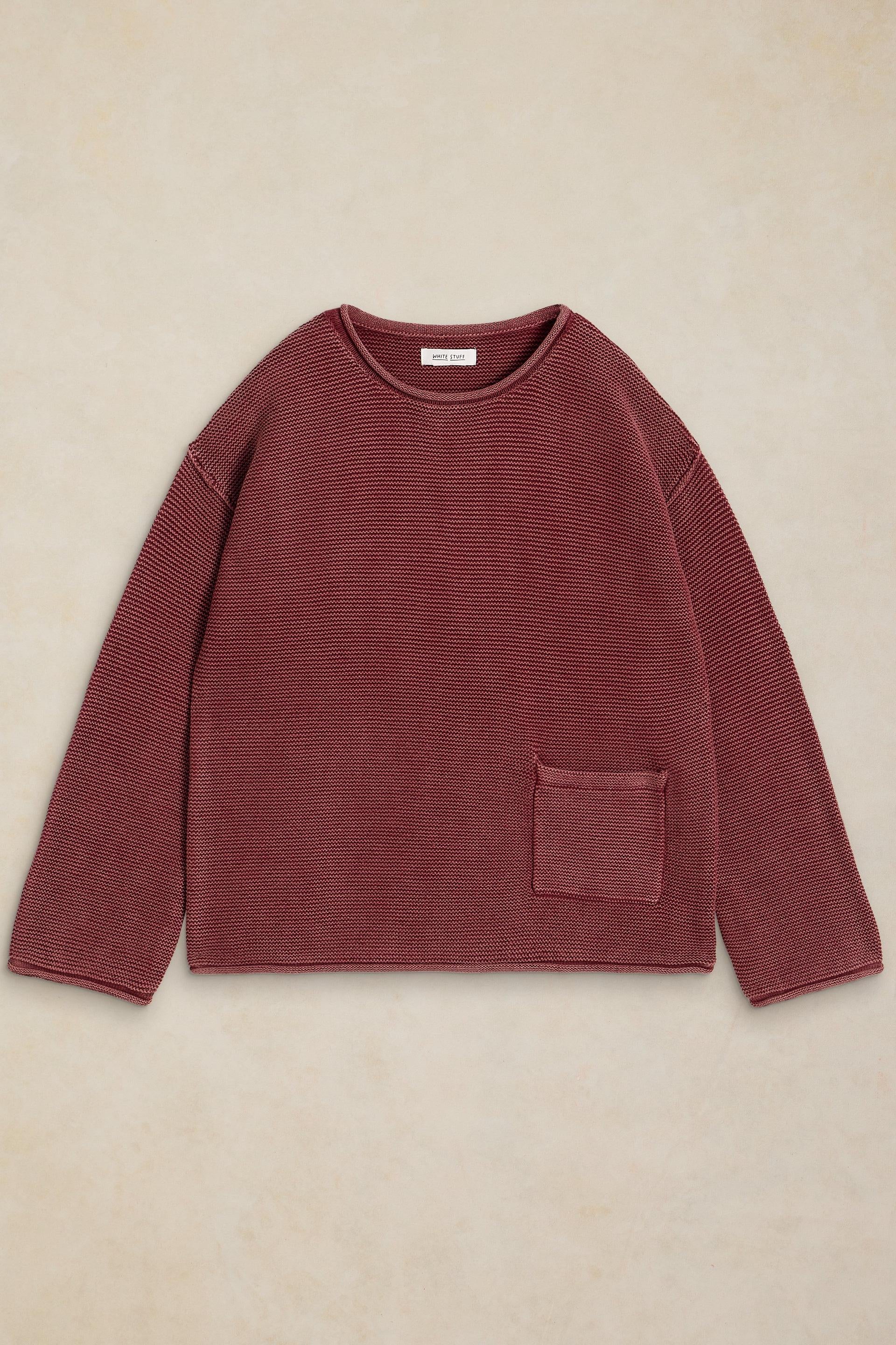 A ribbed knit long-sleeve sweater in a muted burgundy or dusty rose color, laid flat on a textured beige background. It features a crew neck and a single patch pocket on the lower left side.