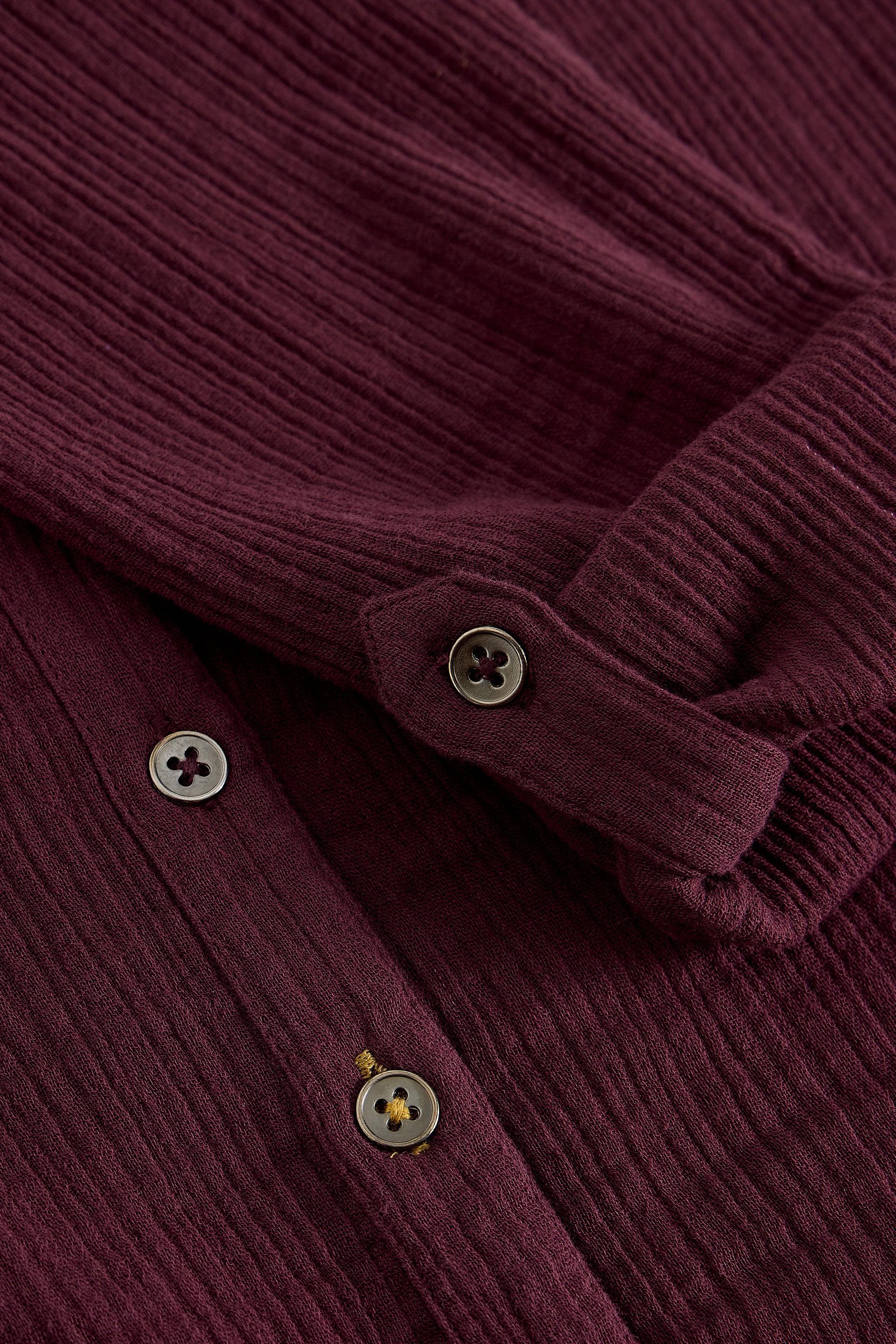 Close-up of a ribbed knit top in deep burgundy, showing the fabric texture, rolled cuff, and metallic buttons.