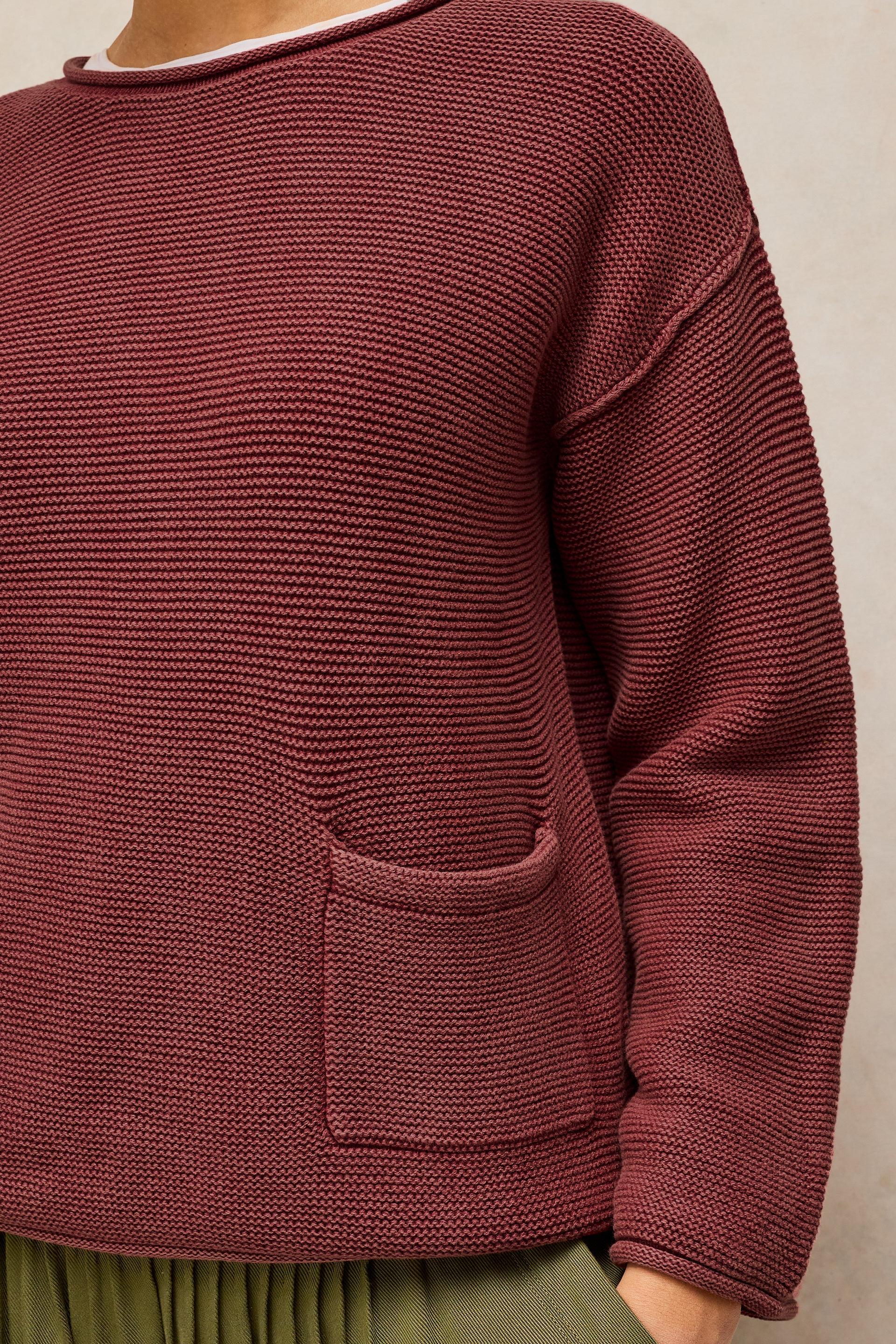 A close-up of a person wearing a ribbed knit sweater in a muted burgundy or maroon shade, paired with an olive green skirt. The sweater features a crew neck, dropped shoulders, and a front patch pocket.