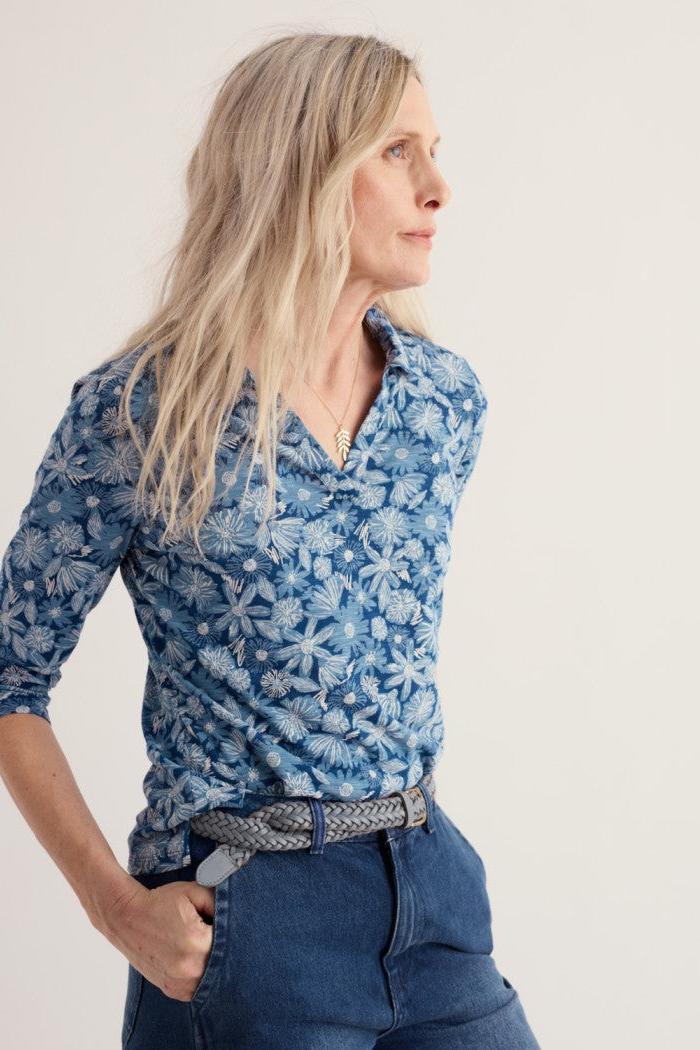 A woman wearing a blue button-down shirt with a white floral pattern, paired with dark blue jeans and a patterned belt, photographed against a plain light background.