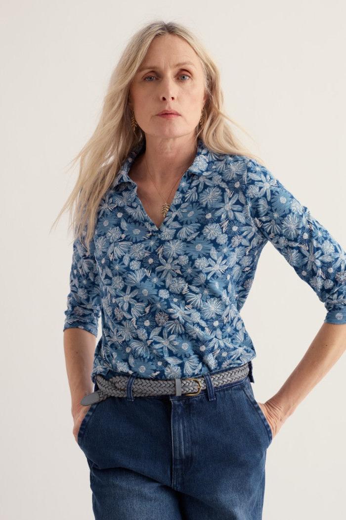 A woman modeling a blue and white floral patterned button-down shirt with three-quarter sleeves, paired with dark blue jeans and a patterned belt, against a plain light background.