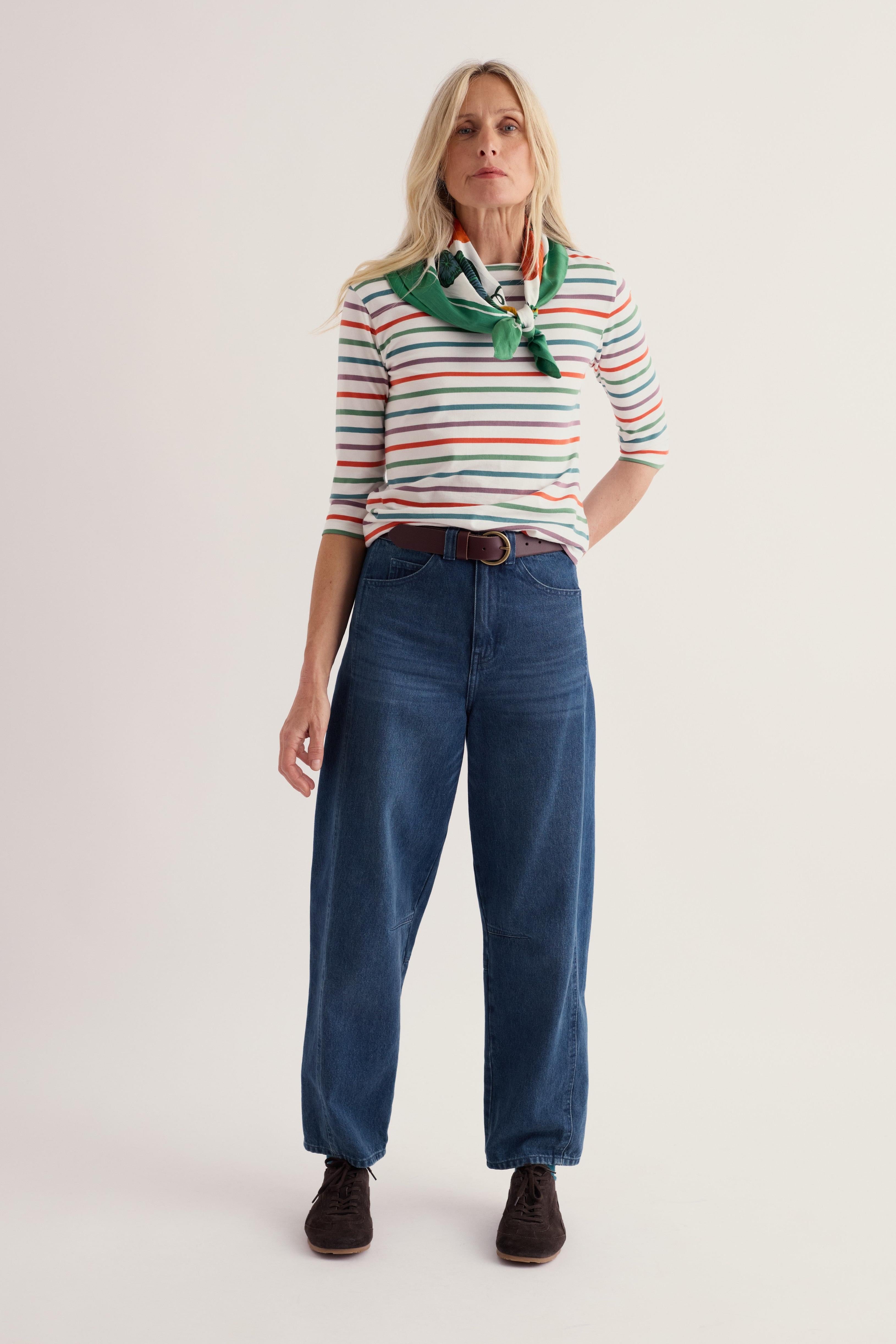 A woman modeling a striped short-sleeve polo shirt with a green neck scarf, paired with dark blue jeans and brown shoes, against a plain light background.