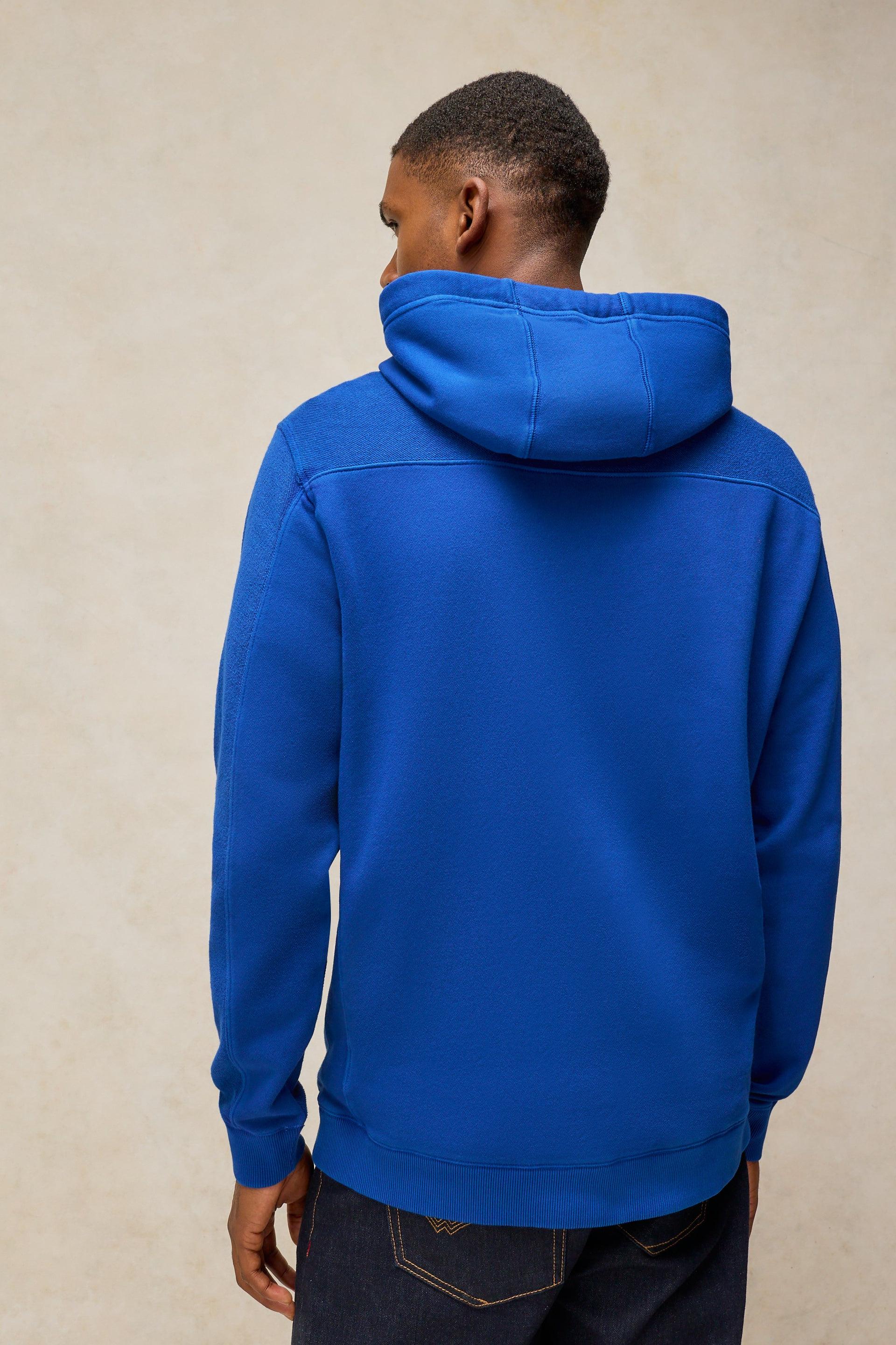 A man viewed from the back wearing a bright royal blue pullover hoodie and dark jeans, standing against a plain beige textured wall.