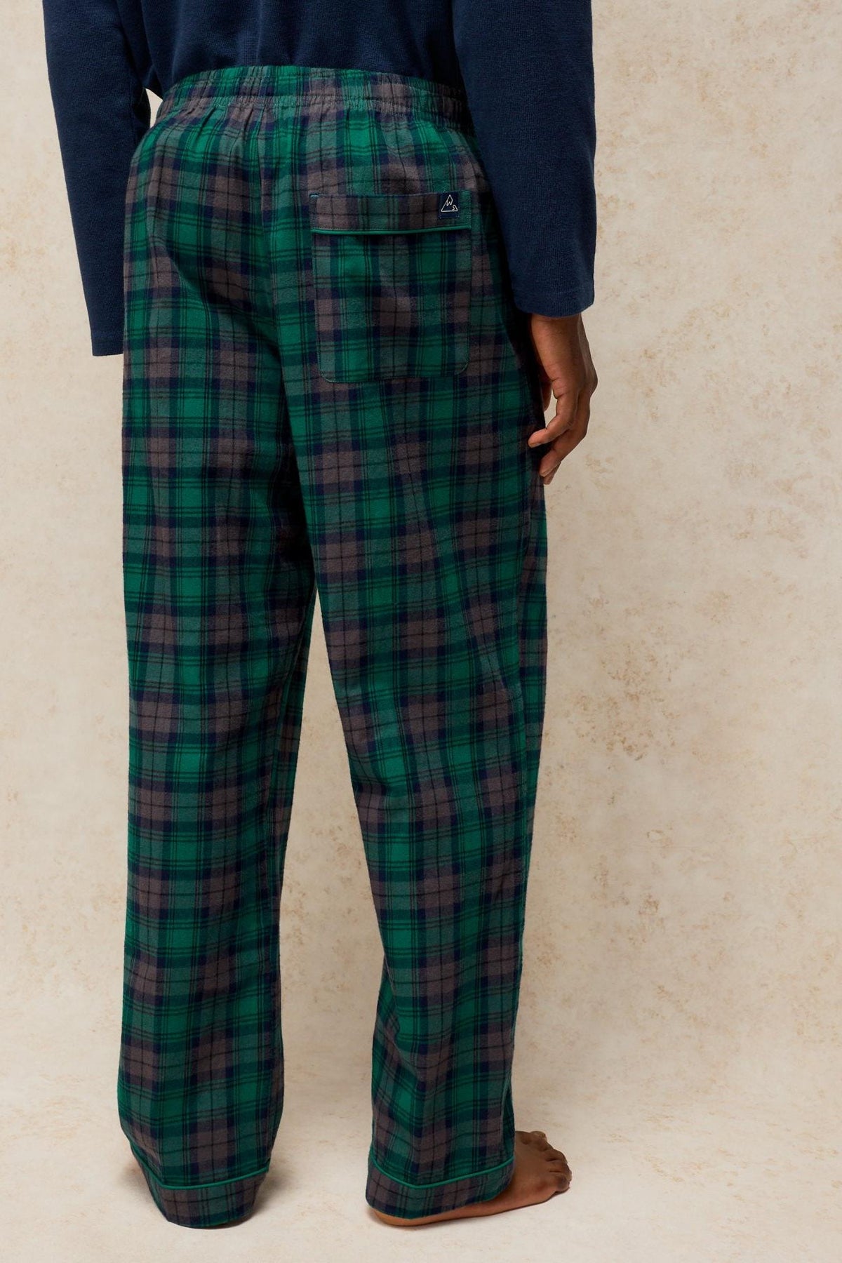 Rear view of a man wearing green plaid White Stuff pyjama trousers with a navy long-sleeve top.