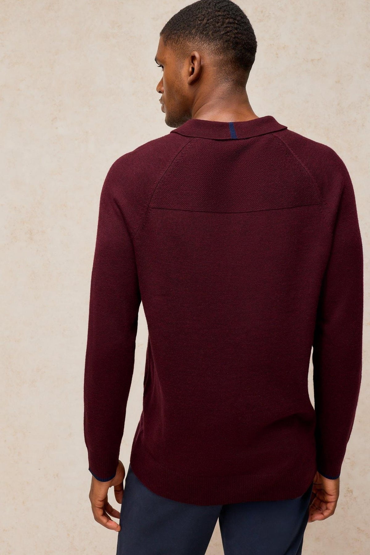 Rear view of a man wearing a plum-coloured White Stuff merino wool long-sleeved polo shirt against a neutral background.