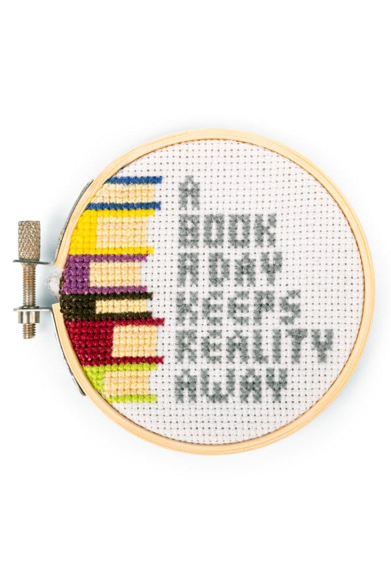 Cross-stitched hoop with 'A book a day keeps reality away' text and colorful book illustrations.