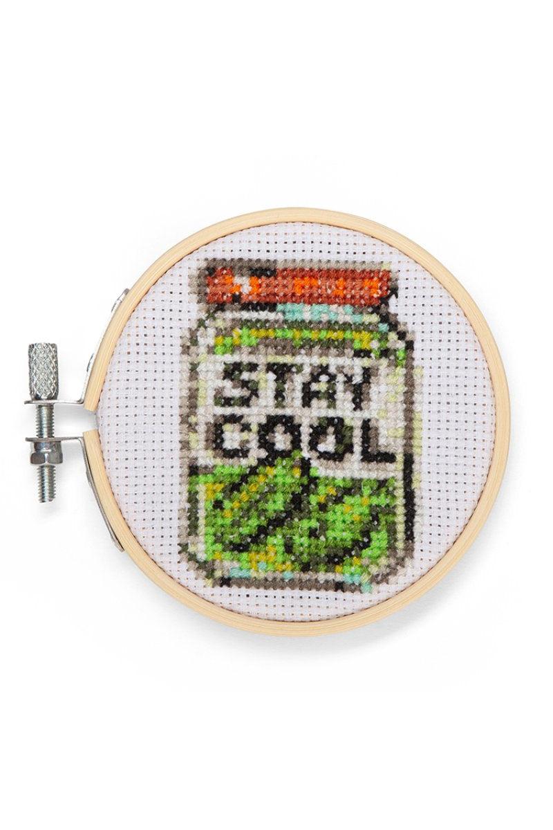 A completed cross-stitch piece inside a wooden embroidery hoop, depicting a canister with the words 'STAY COOL' in black letters, with green and yellow striped lower section and an orange-red top, shown on a white background.