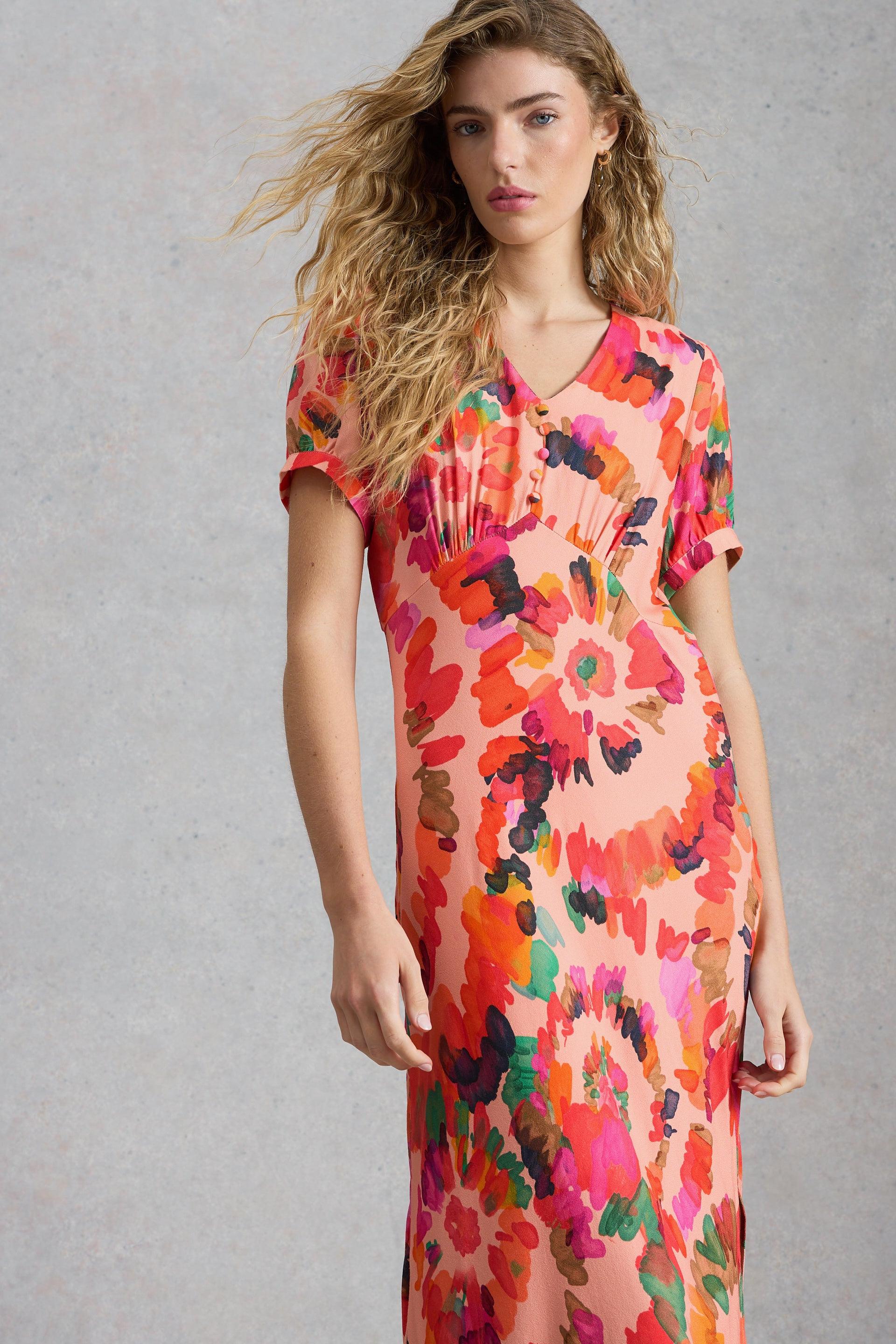 A model with wavy blonde hair wearing a bright peach floral occasion dress against a grey background.