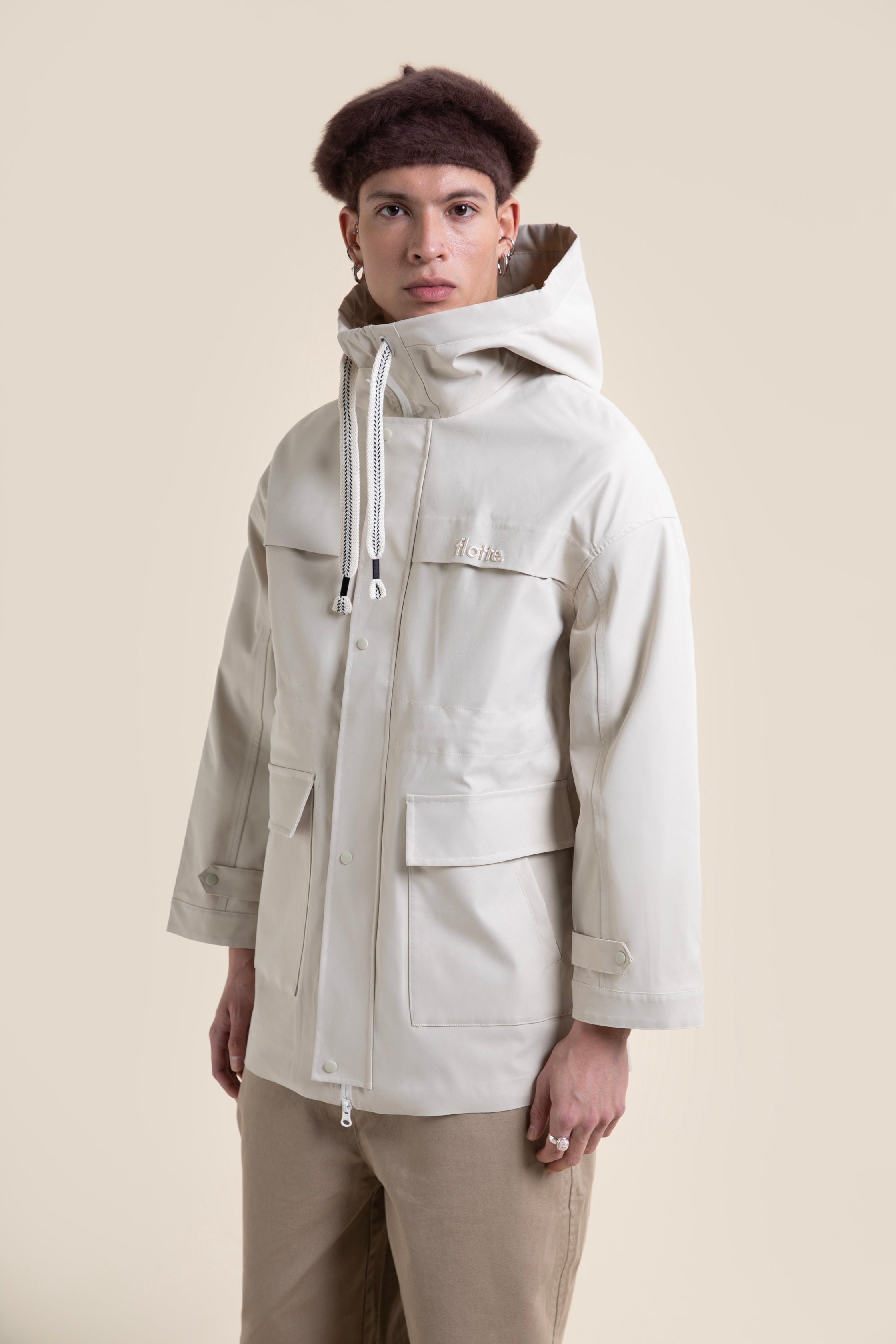 A model wearing a beige Flotte waterproof parka coat with a hood against a light background.