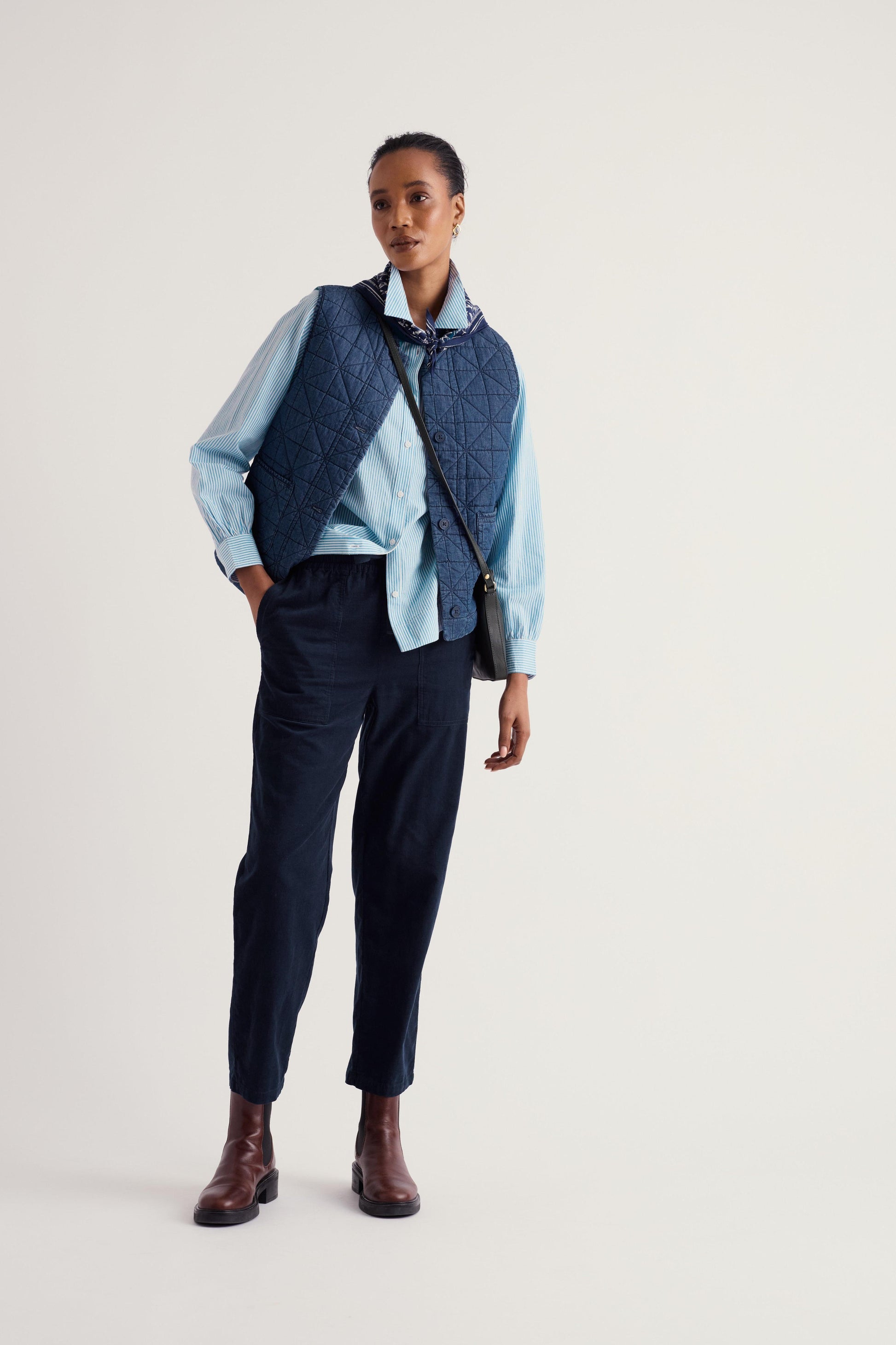 A model stands against a light gray studio background wearing a light blue button-up shirt, a dark navy quilted vest, dark navy trousers, brown leather ankle boots, and a small black crossbody bag.