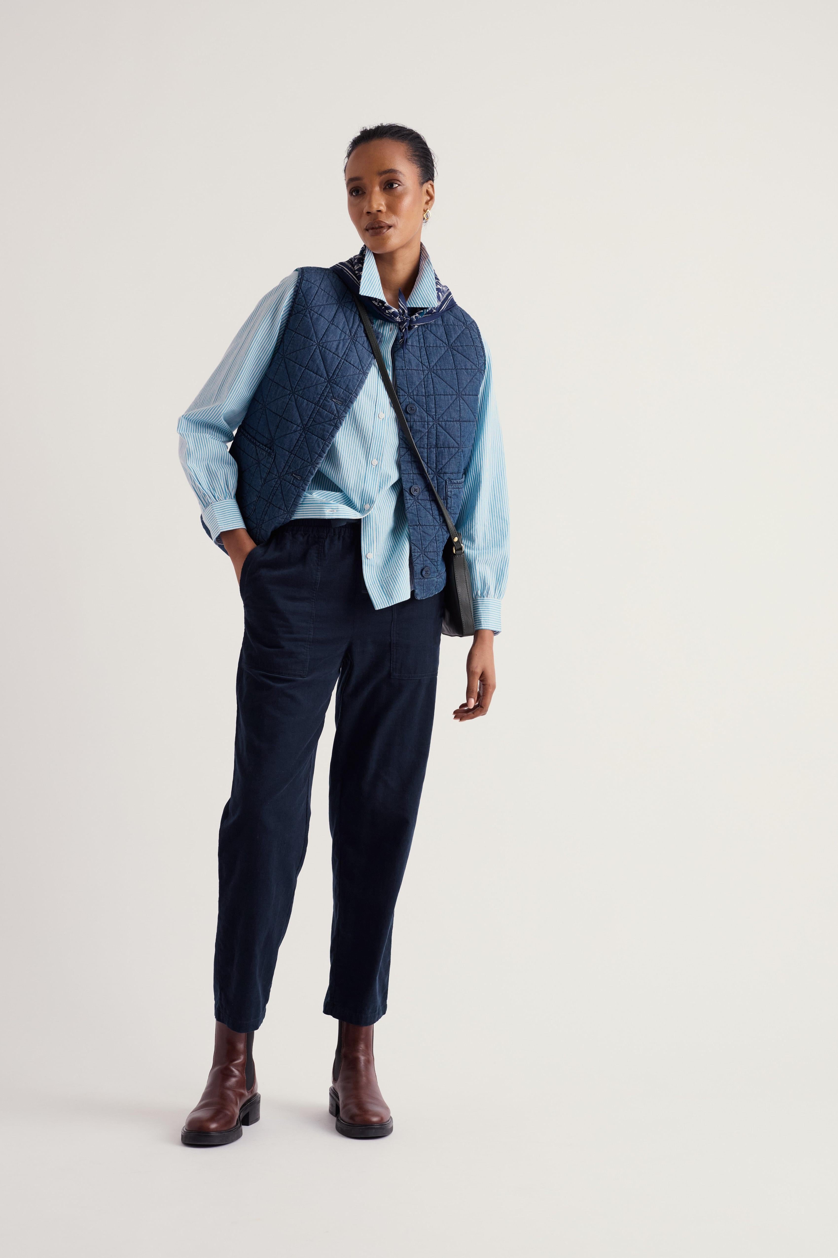 A model stands against a light gray studio background wearing a light blue button-up shirt, a dark navy quilted vest, dark navy trousers, brown leather ankle boots, and a small black crossbody bag.