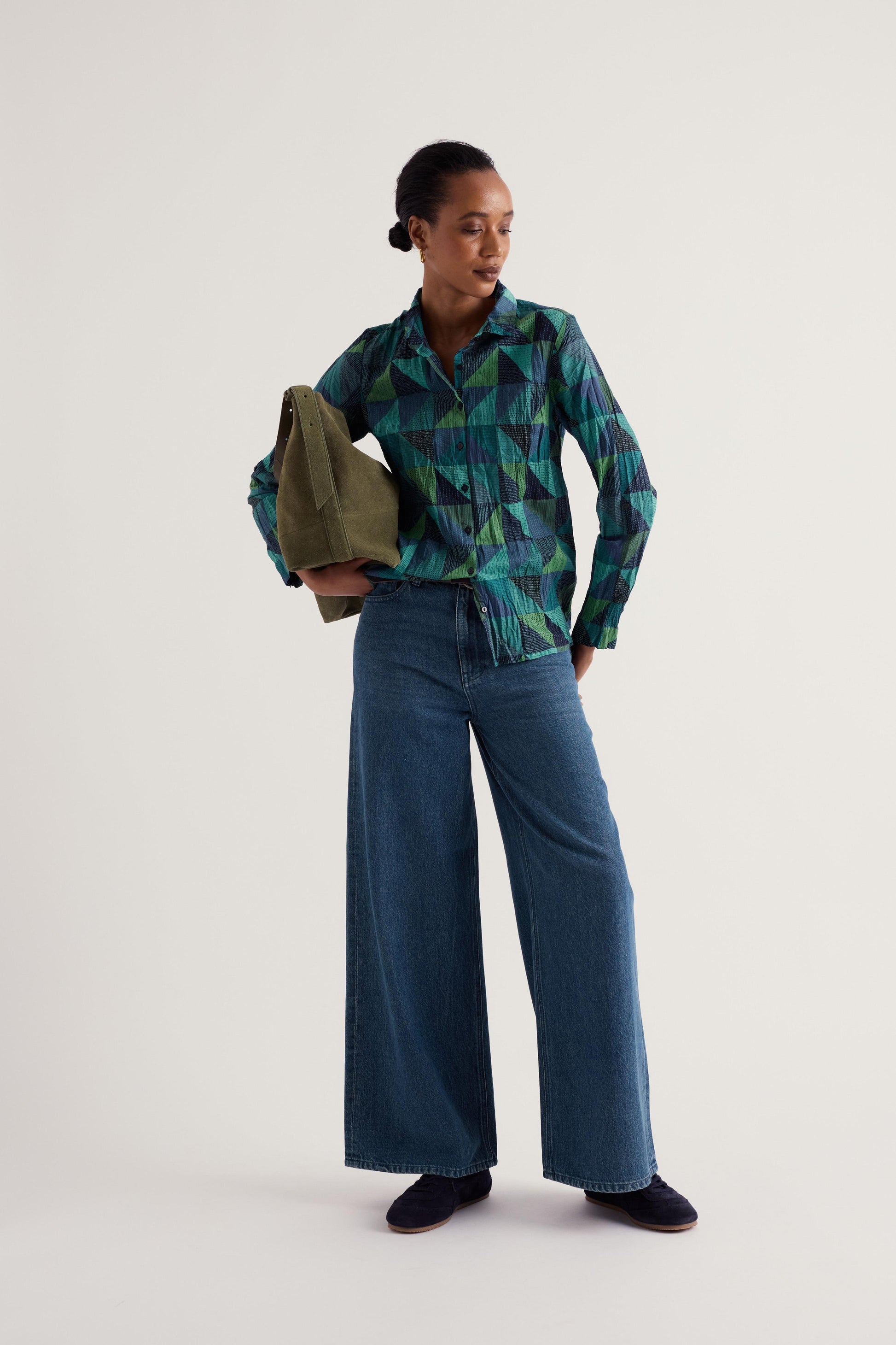 A model stands against a plain light gray studio background wearing a green and blue geometric patterned button-up shirt, wide-leg blue jeans, dark slip-on shoes, and holding an olive green tote bag.