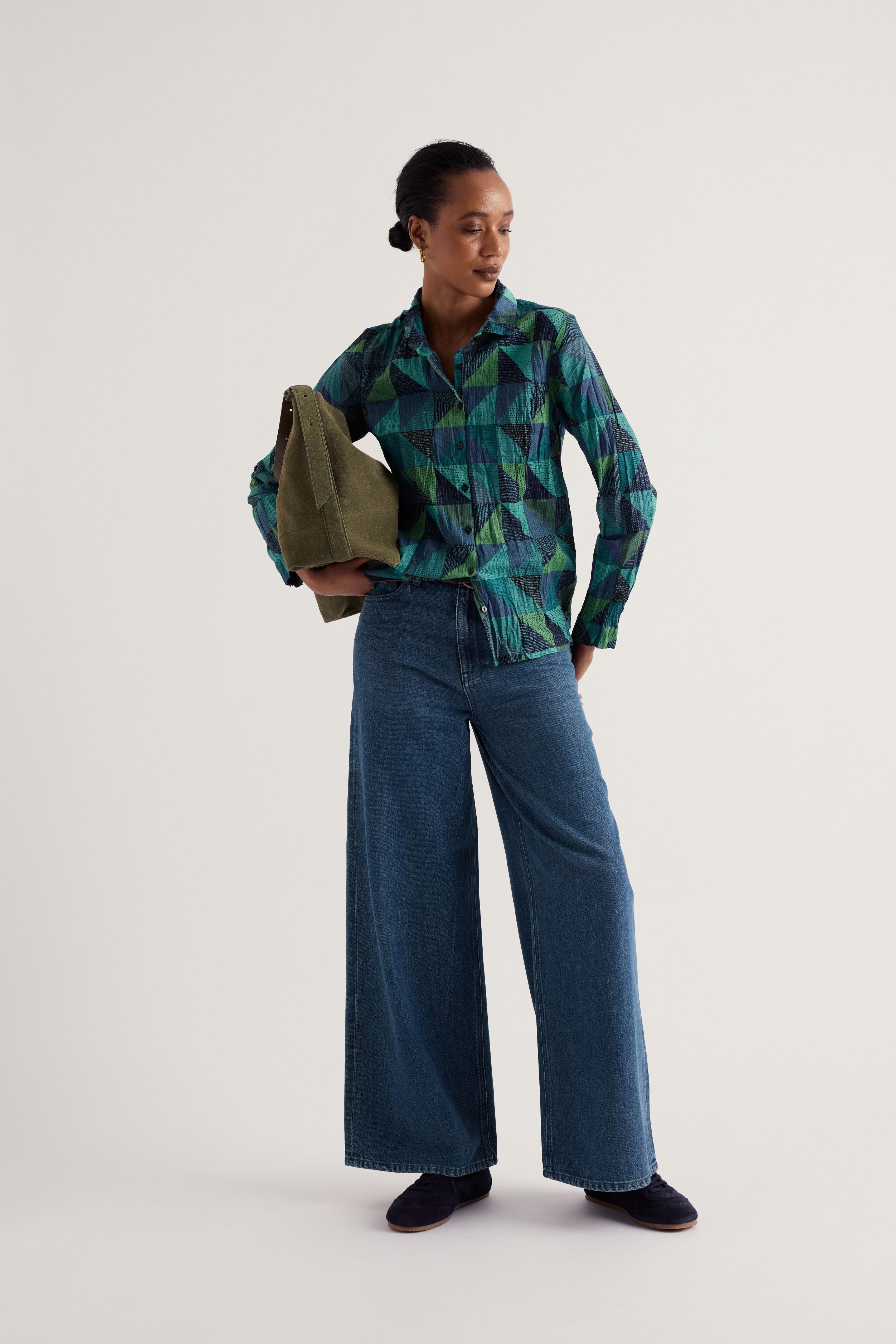A model stands against a plain light gray studio background wearing a green and blue geometric patterned button-up shirt, wide-leg blue jeans, dark slip-on shoes, and holding an olive green tote bag.