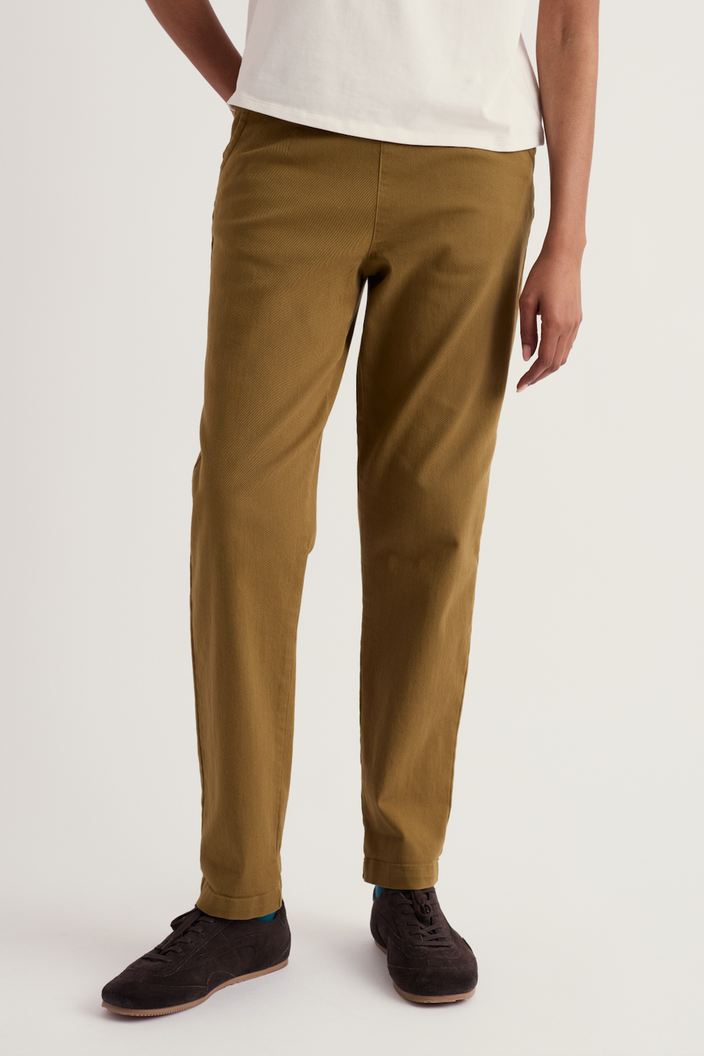 A person wearing slim-fit olive-brown chinos with a white t-shirt and dark brown suede sneakers, shown from the waist down on a light gray studio background.