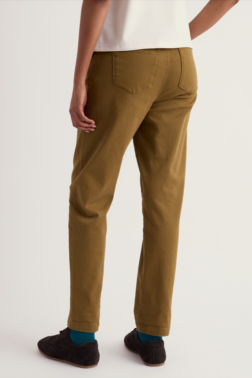 A person shown from the back wearing slim-fit trousers in a warm camel-brown color, paired with a white top, teal socks, and dark slip-on shoes against a plain light background.