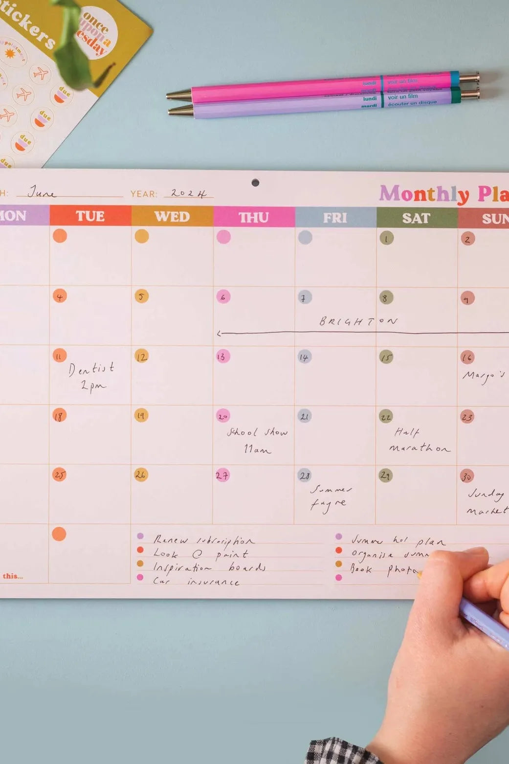 A hand writing on a colorful monthly wall calendar tear-off pad with pens and stickers nearby.