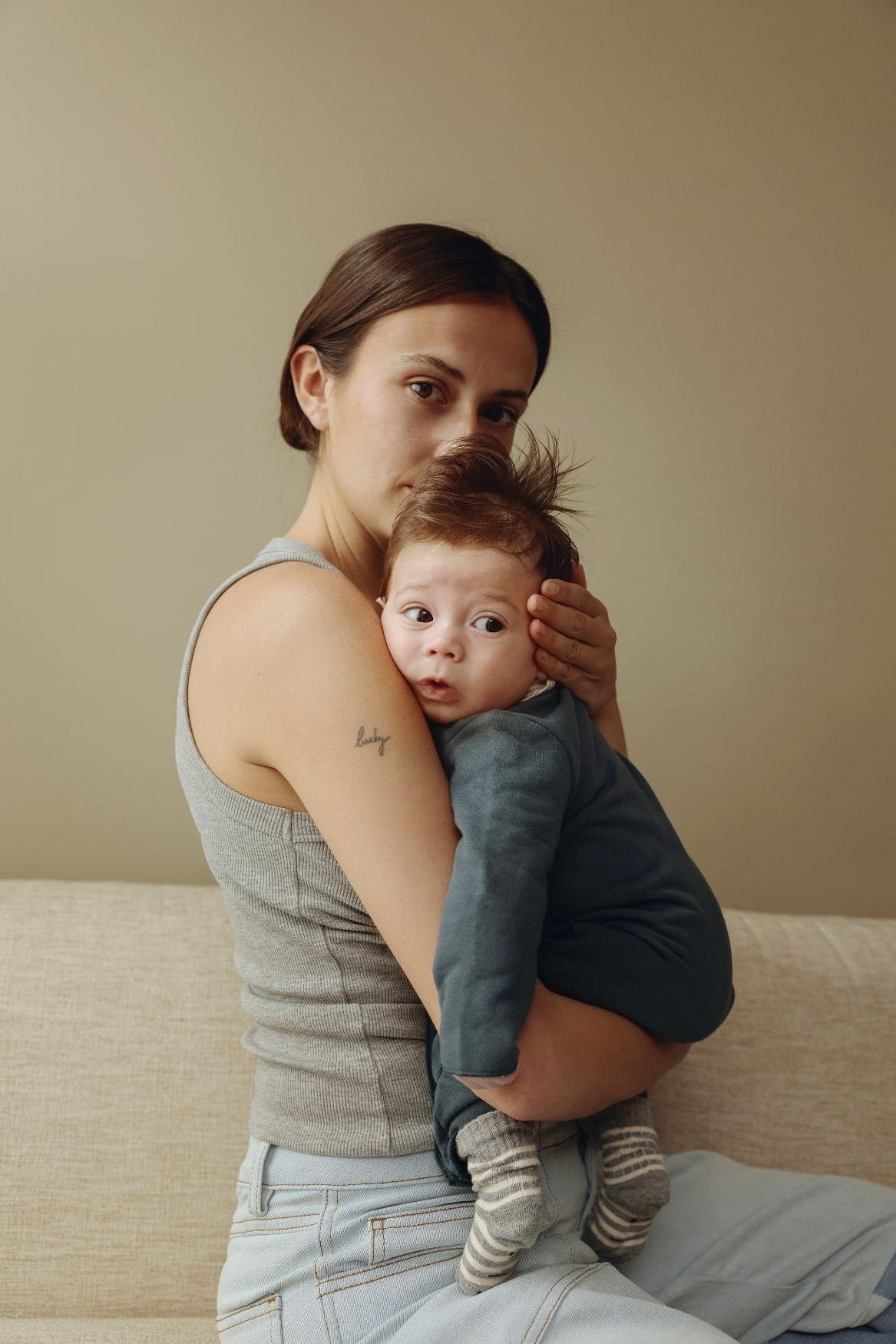 A woman sitting on a beige sofa holds a baby. The woman wears a gray sleeveless top and light blue jeans, and the baby is dressed in a dark blue outfit with striped socks.