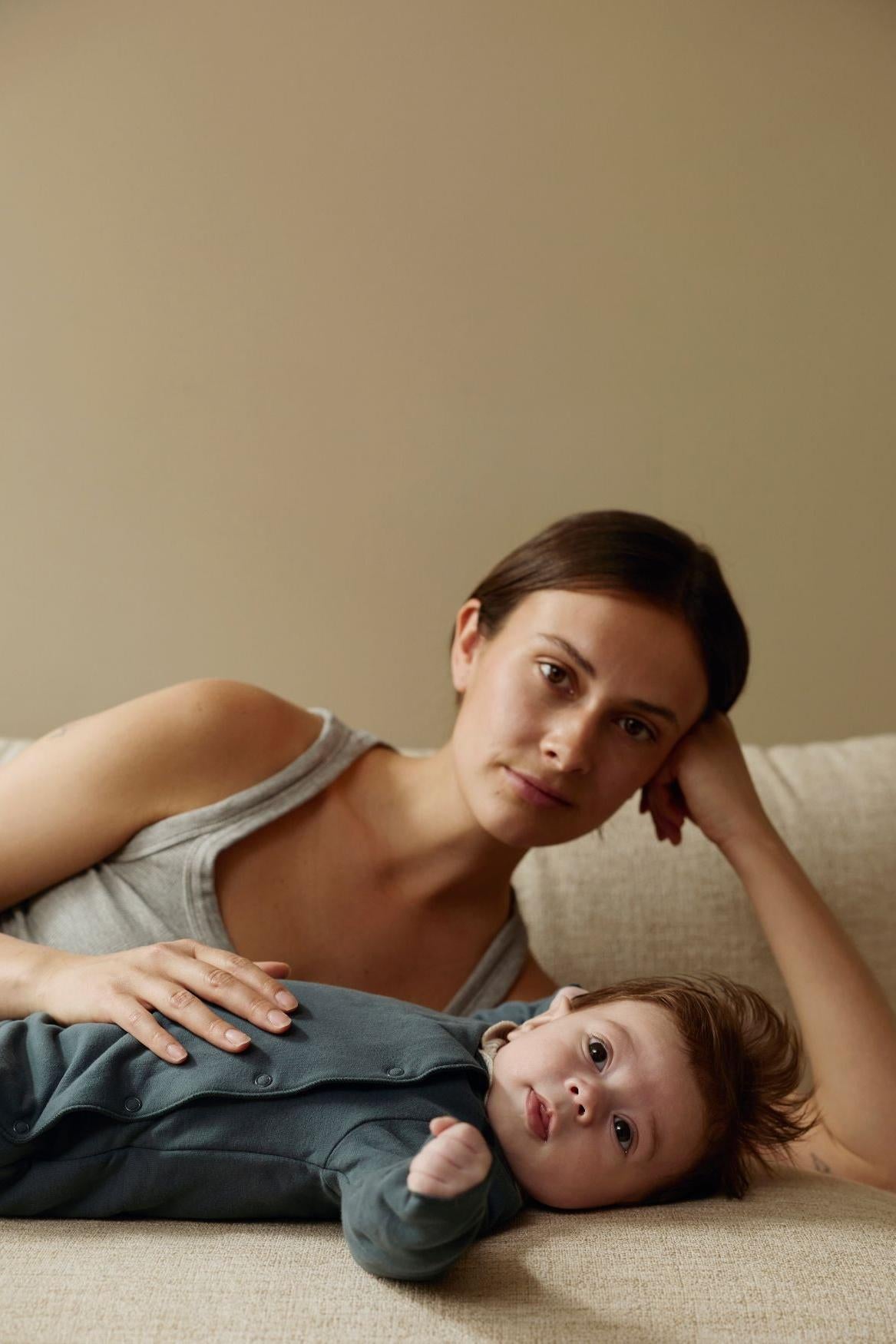 A woman in a gray tank top lies on a beige couch with a baby wearing a dark blue-gray long-sleeve romper. The background is a solid beige wall.