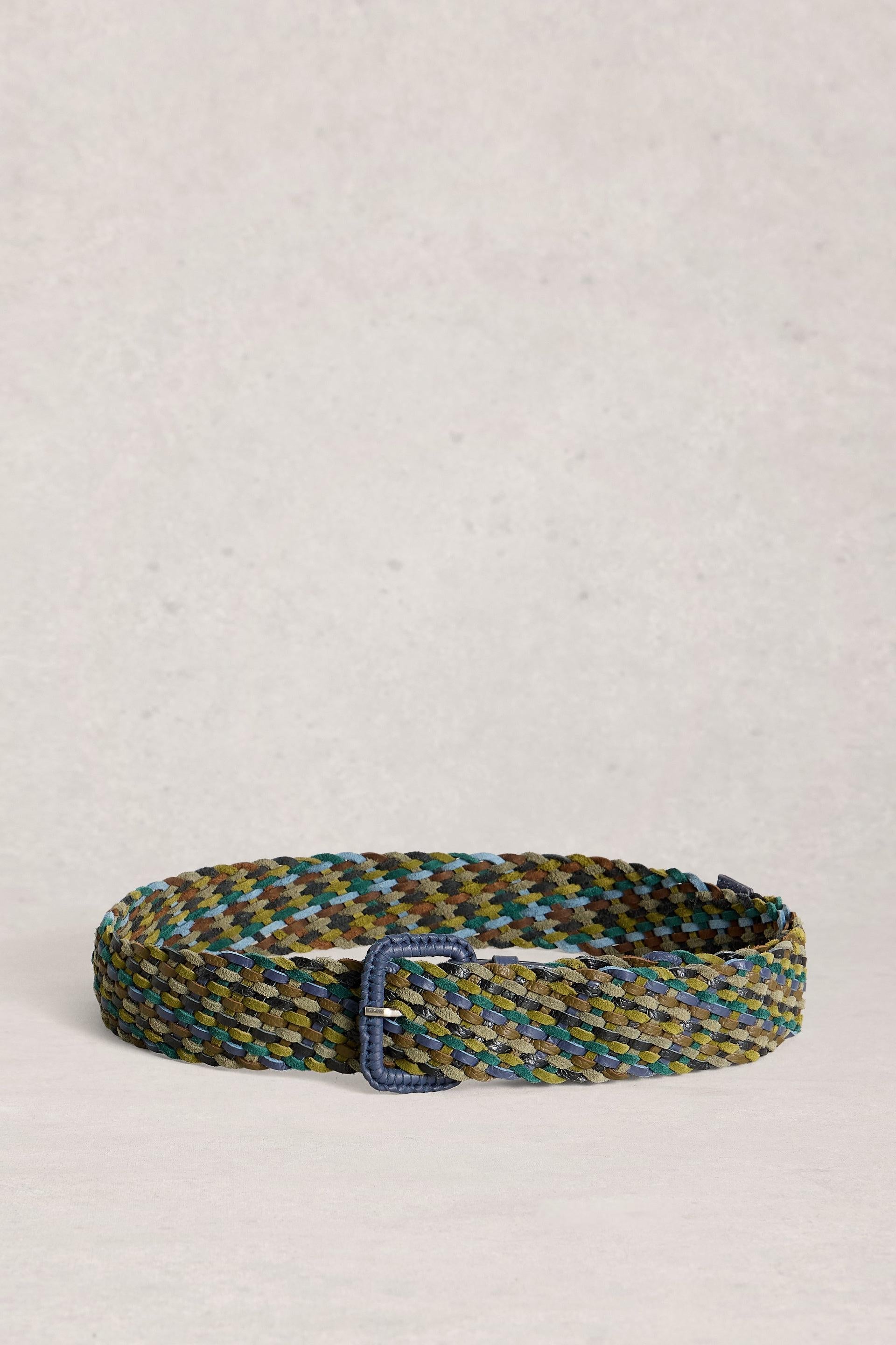 A woven belt with a multicolor pattern of olive green, blue, and gold, featuring a dark blue buckle, displayed on a light beige textured background.