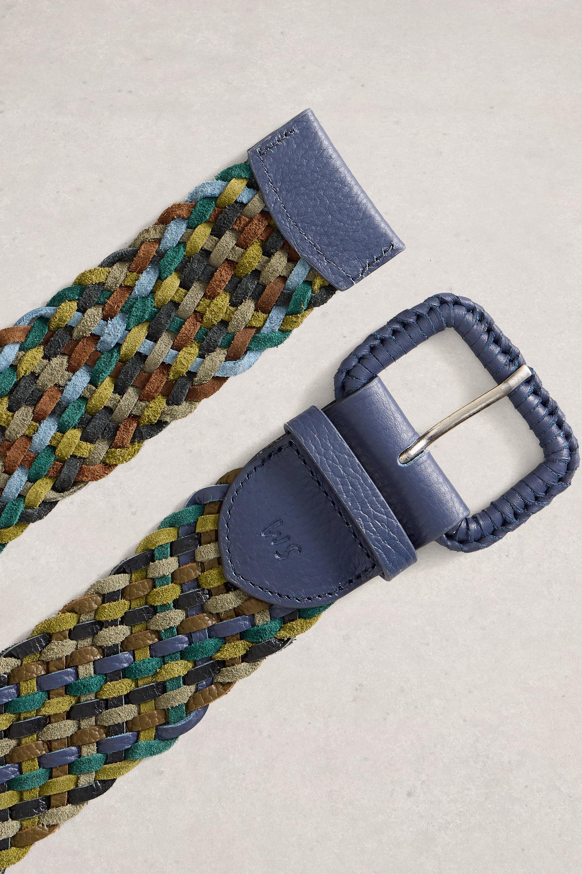 A woven belt with a multicolor braided strap in shades of green, yellow, blue, and brown, finished with a navy blue leather buckle and keeper, shown on a light beige textured background.