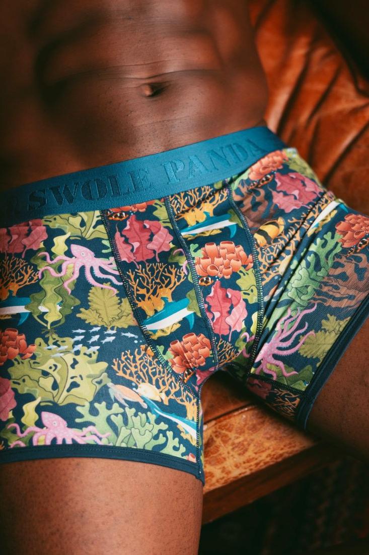 A muscular man wearing colorful ocean-print bamboo boxer briefs while sitting in a leather chair.