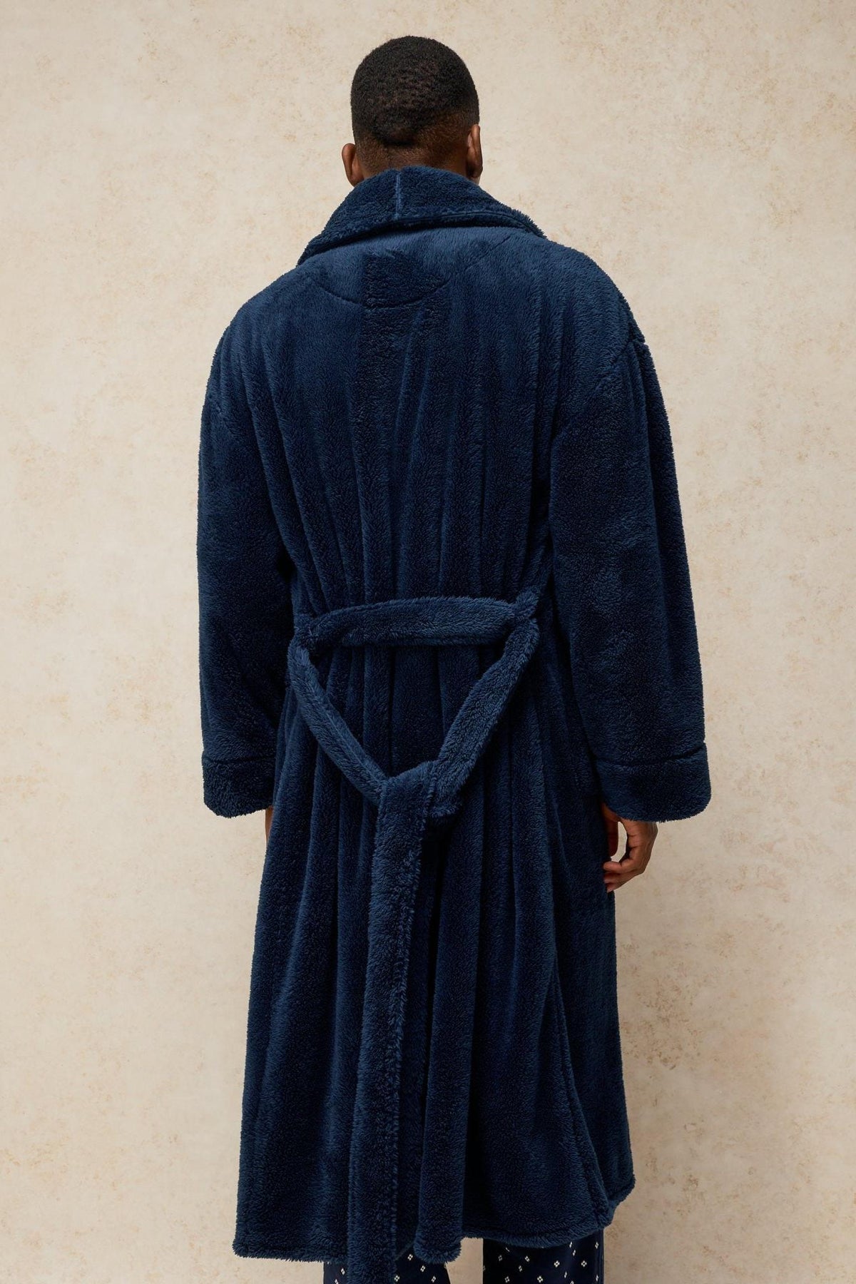 Rear view of a man wearing a cozy navy fluffy White Stuff dressing gown against a neutral background.