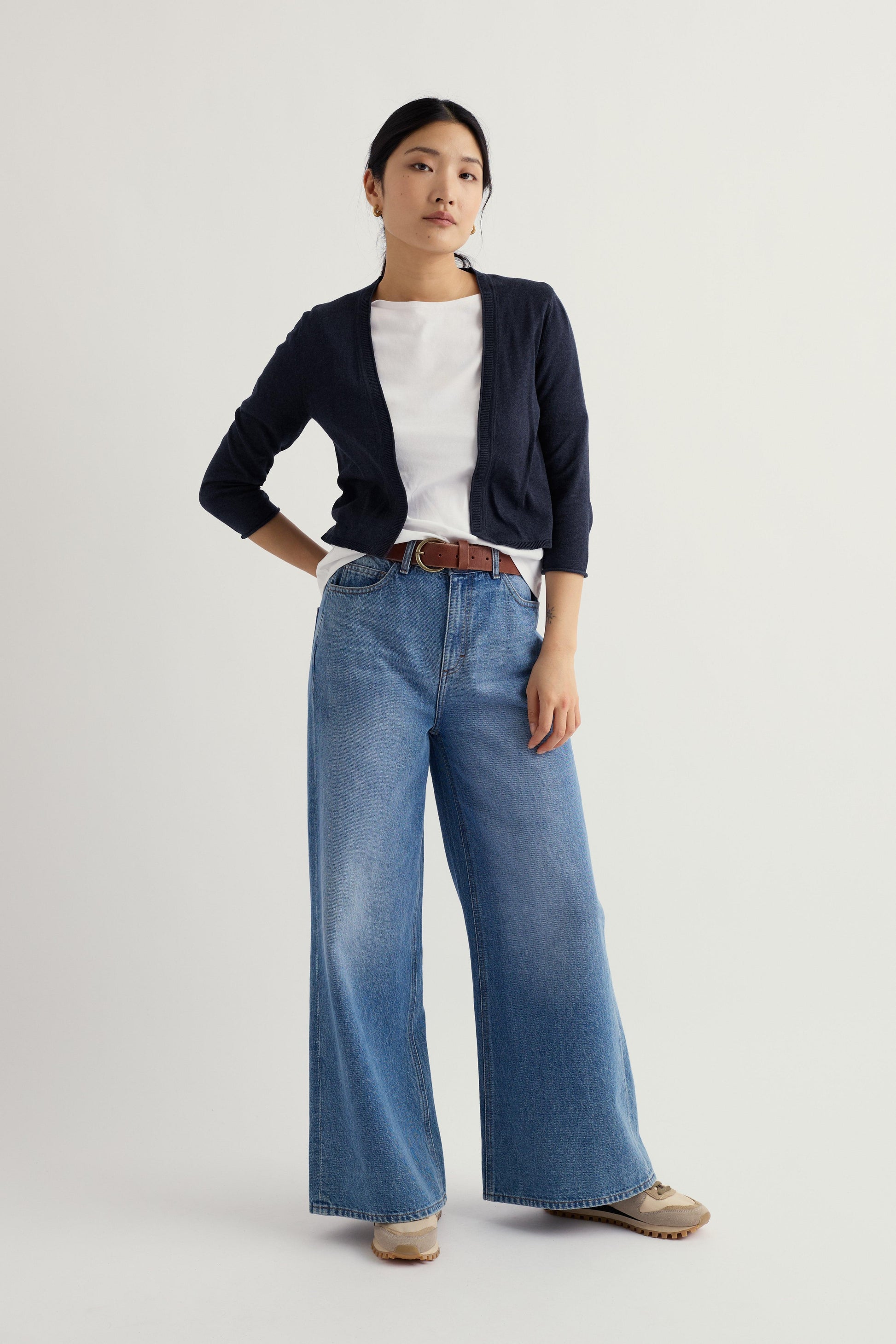 Woman wearing a navy Seasalt cardigan, white shirt, and blue jeans on a light gray background