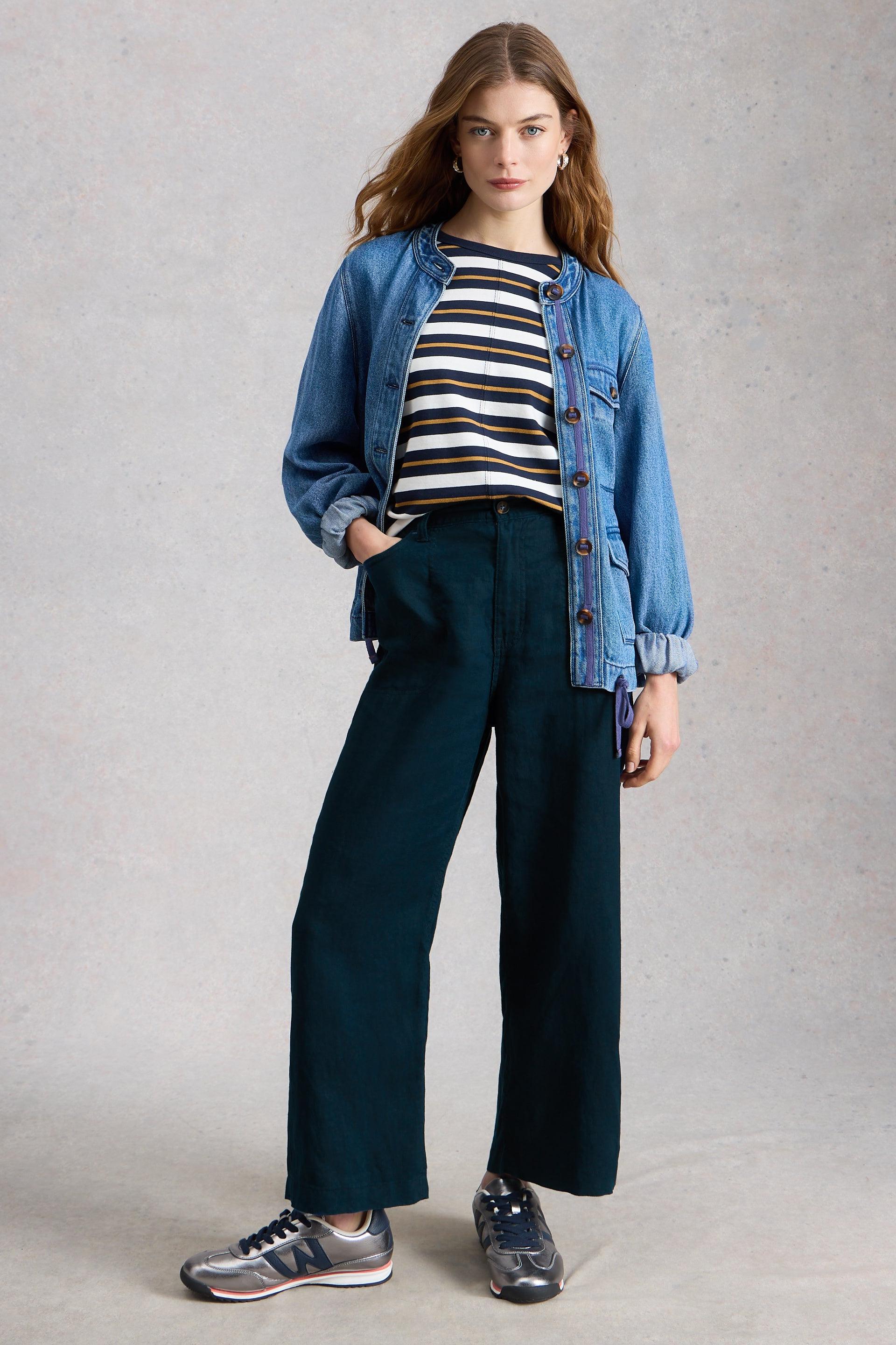 Woman wearing a striped shirt, denim jacket, and wide-leg trousers on a plain background