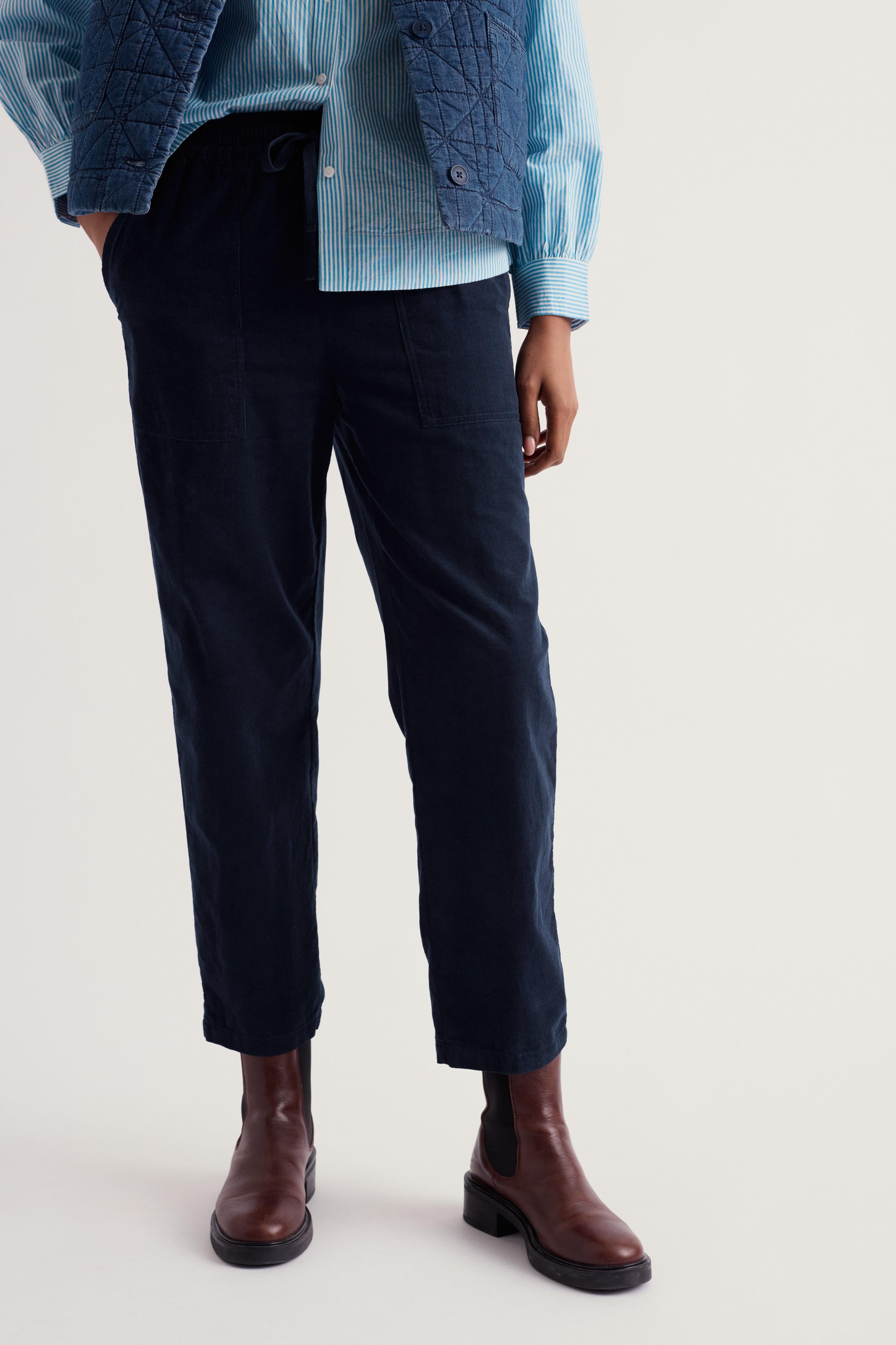 A person wearing dark navy linen trousers, styled with a light blue striped shirt, a blue quilted vest, and brown leather Chelsea boots, photographed against a plain light gray studio background.