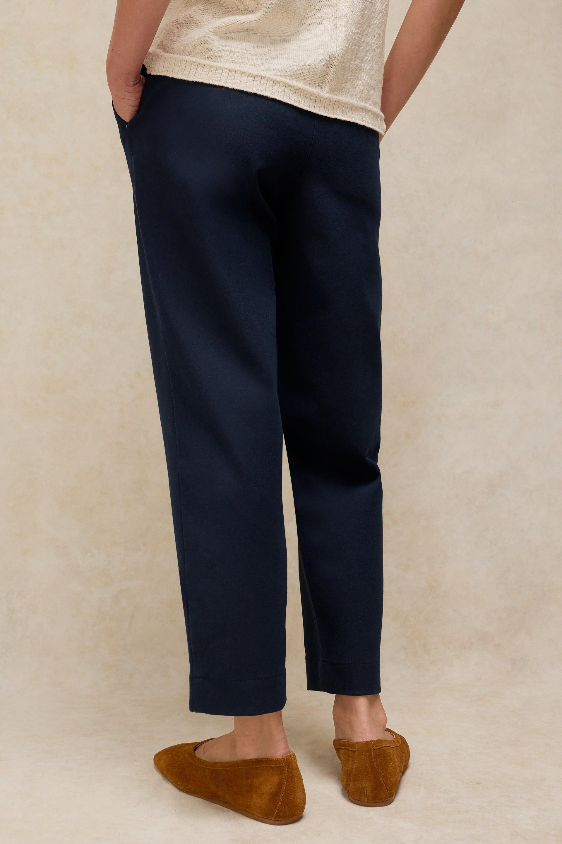 A person shown from the waist down wearing dark navy cropped trousers, a cream knit top, and brown suede flats against a textured beige studio backdrop.
