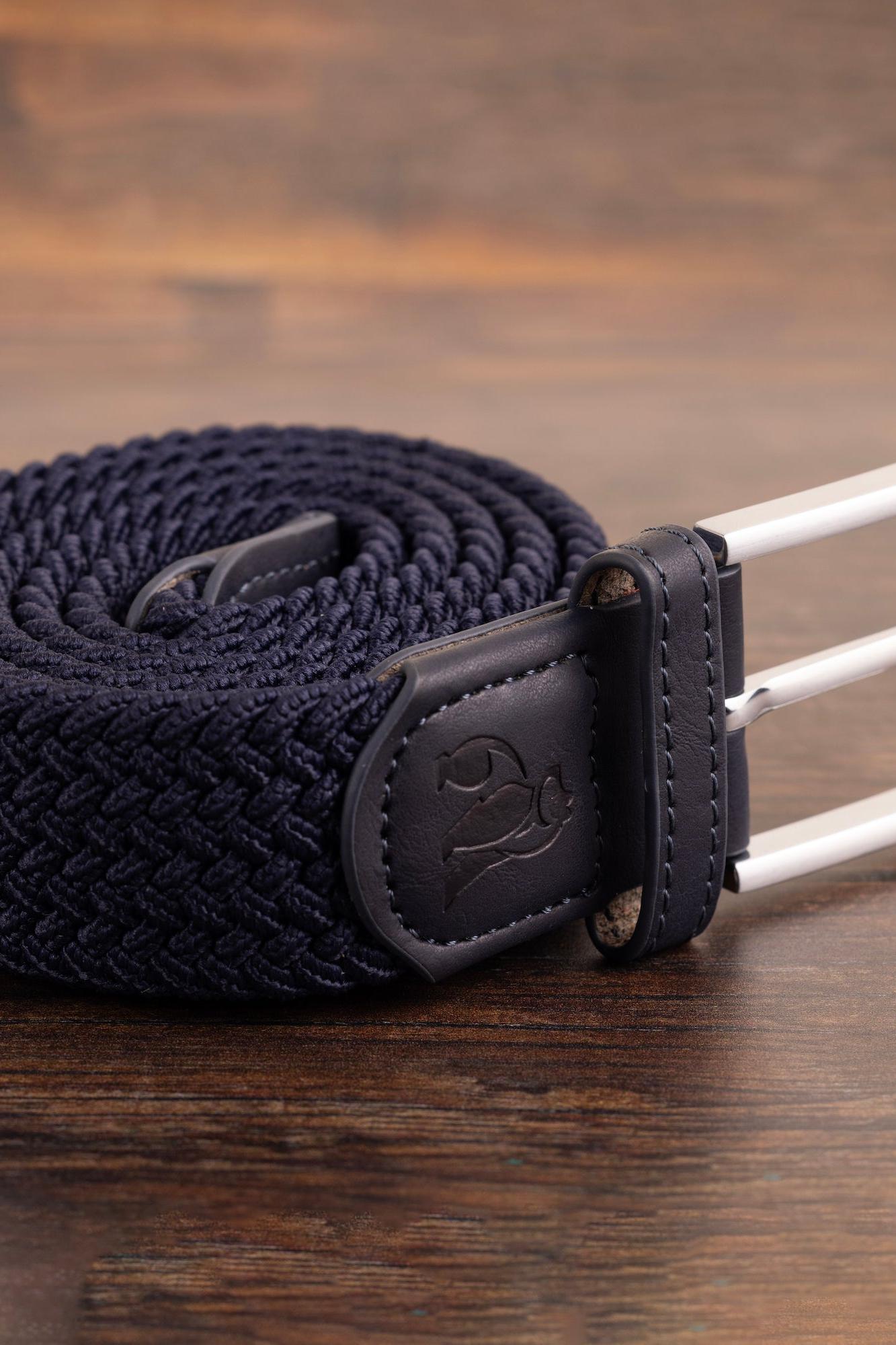 Coiled navy woven belt with leather trim and a silver buckle resting on a wooden background.