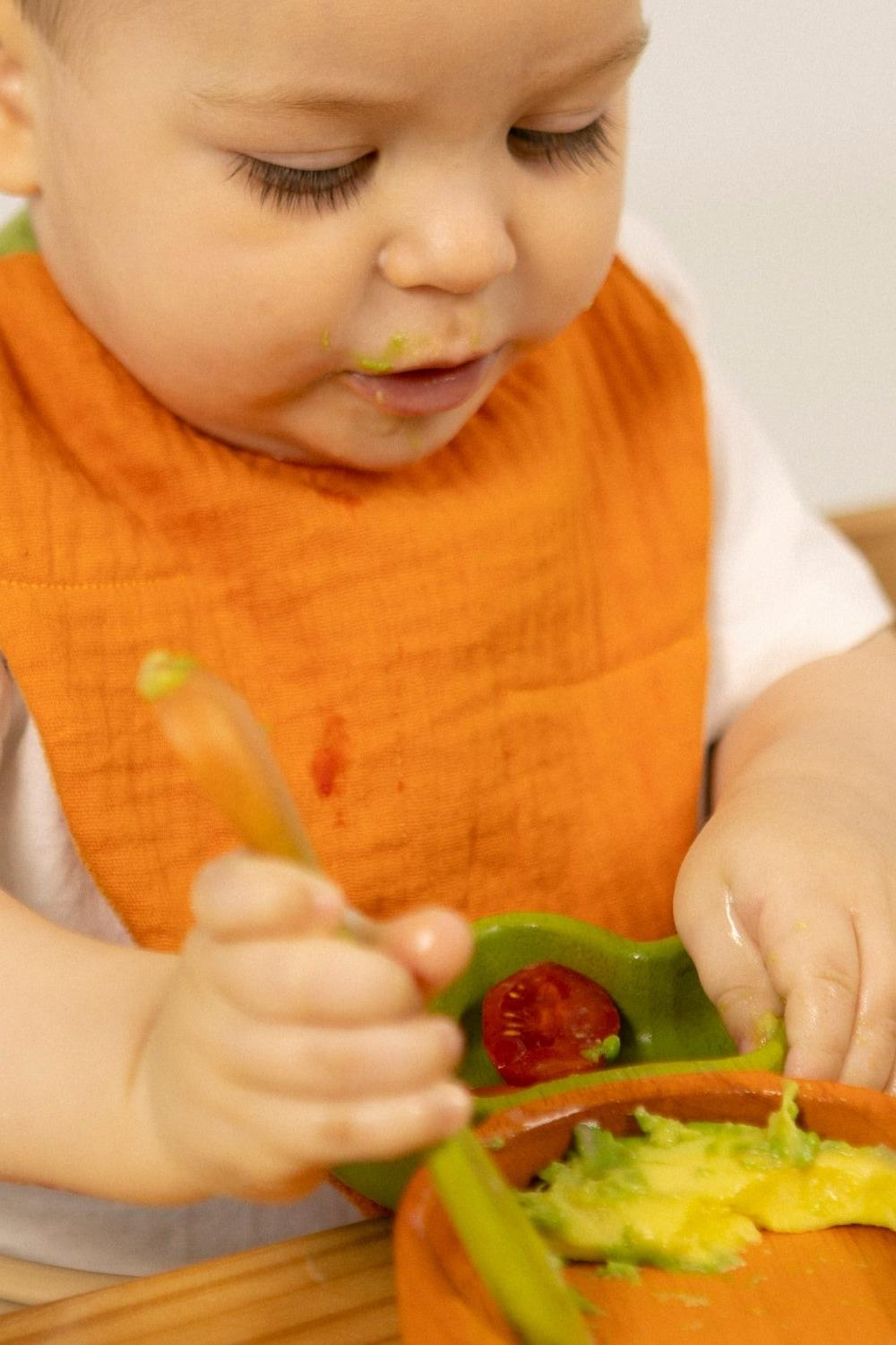 Baby in an orange Oli&Carol bib eating with a spoon and fork