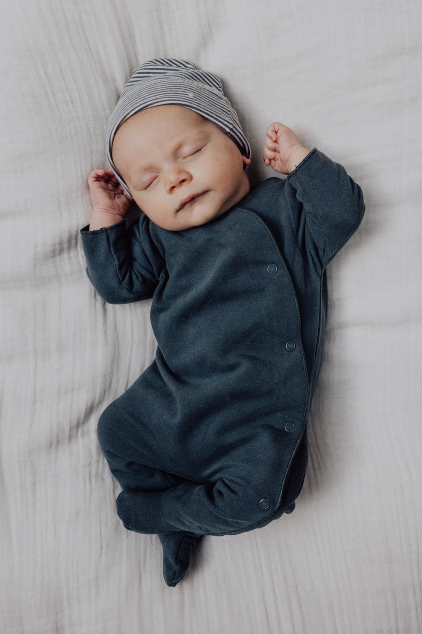 Baby sleeping peacefully wearing a dark blue outfit and striped hat on a soft surface.