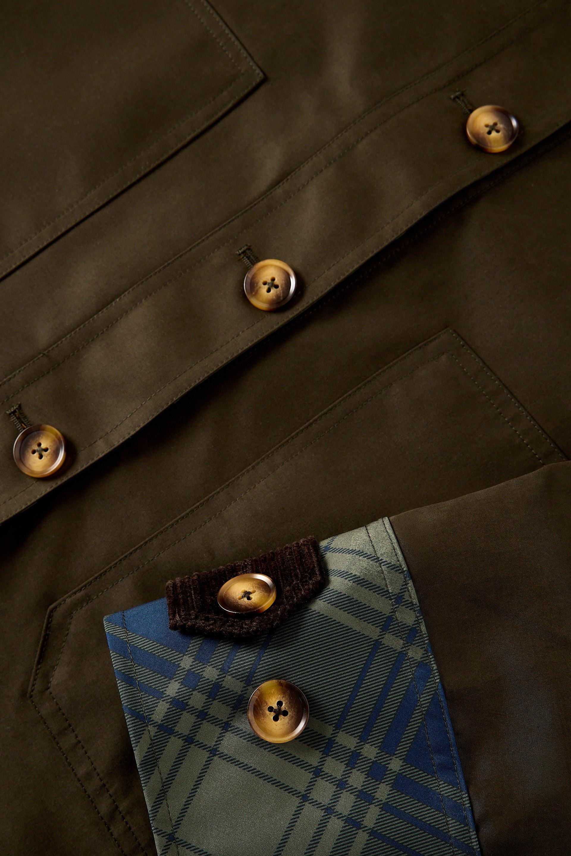 Close-up detail of an olive brown waxed cotton jacket, showing the buttoned flap collar with a blue and green plaid shirt underneath and brass buttons.