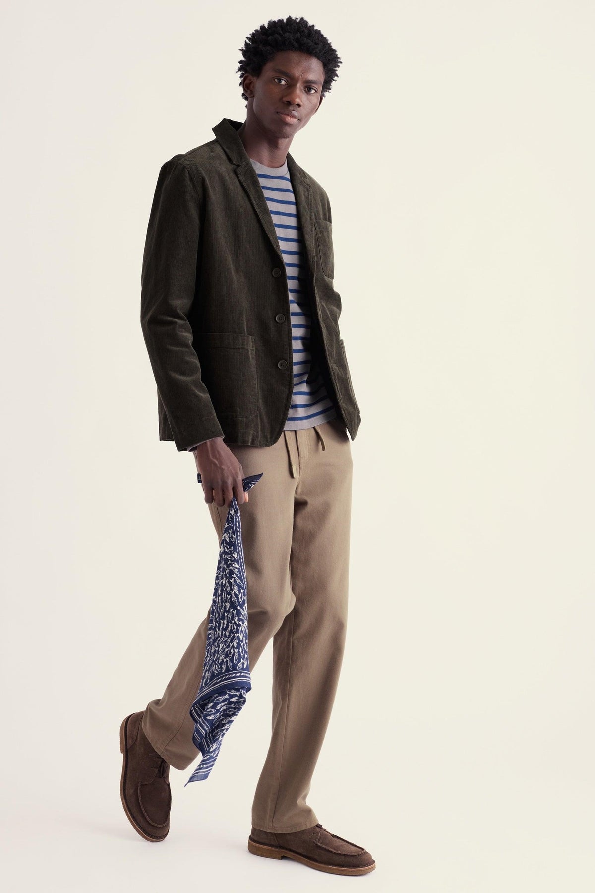 A man wearing an olive green corduroy blazer over a striped shirt and tan trousers.