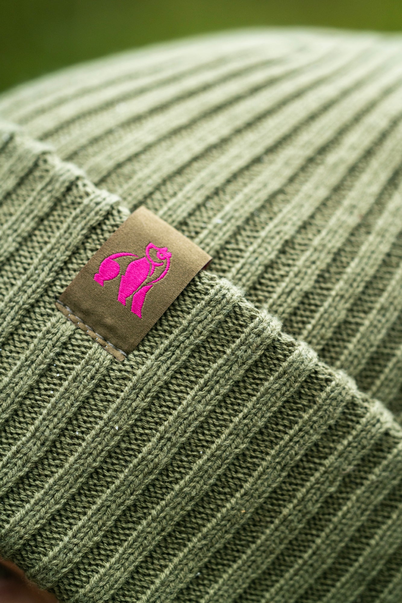 Green organic cotton knit beanie with a pink logo patch on a blurred green background