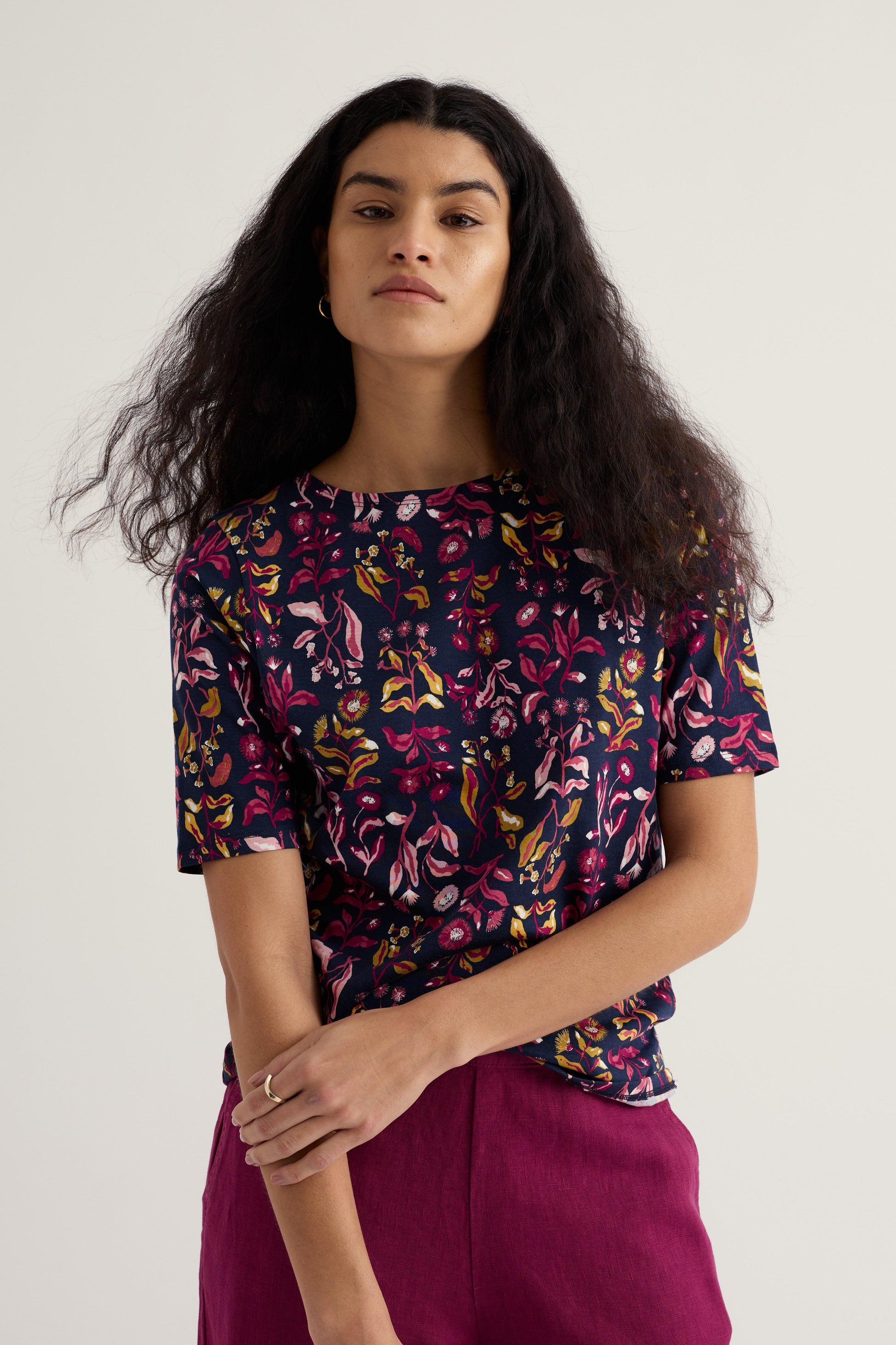 Woman wearing a colorful patterned top and pink pants against a plain background
