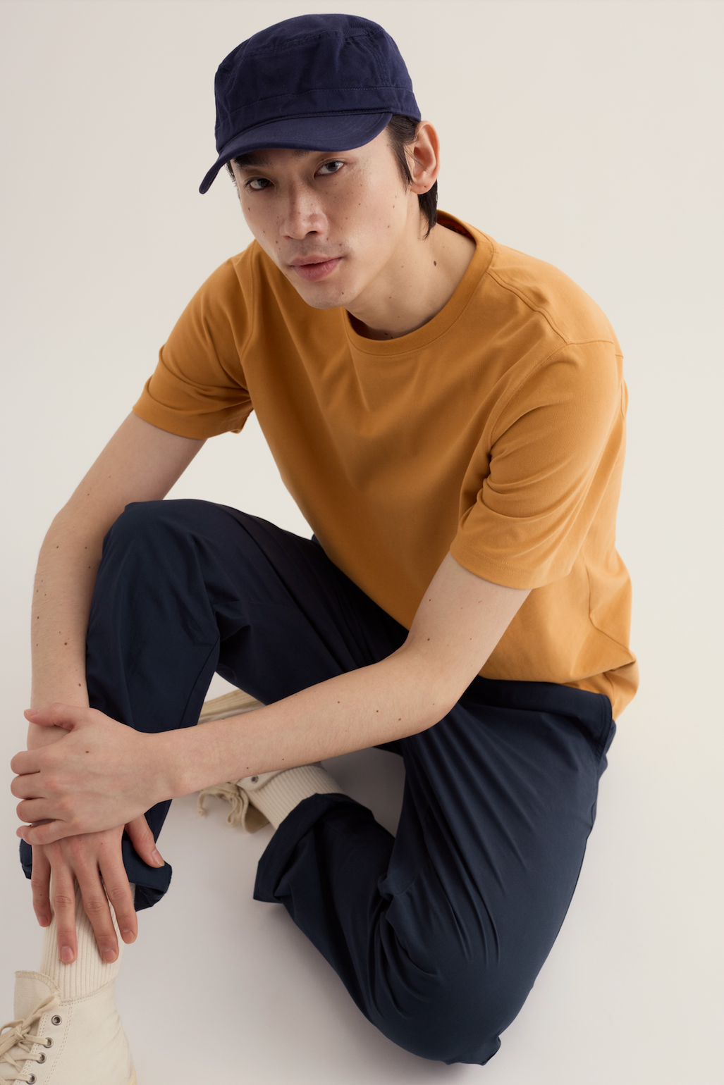 A man wearing a mustard organic cotton men's t-shirt and navy baseball cap sitting on a plain studio floor.