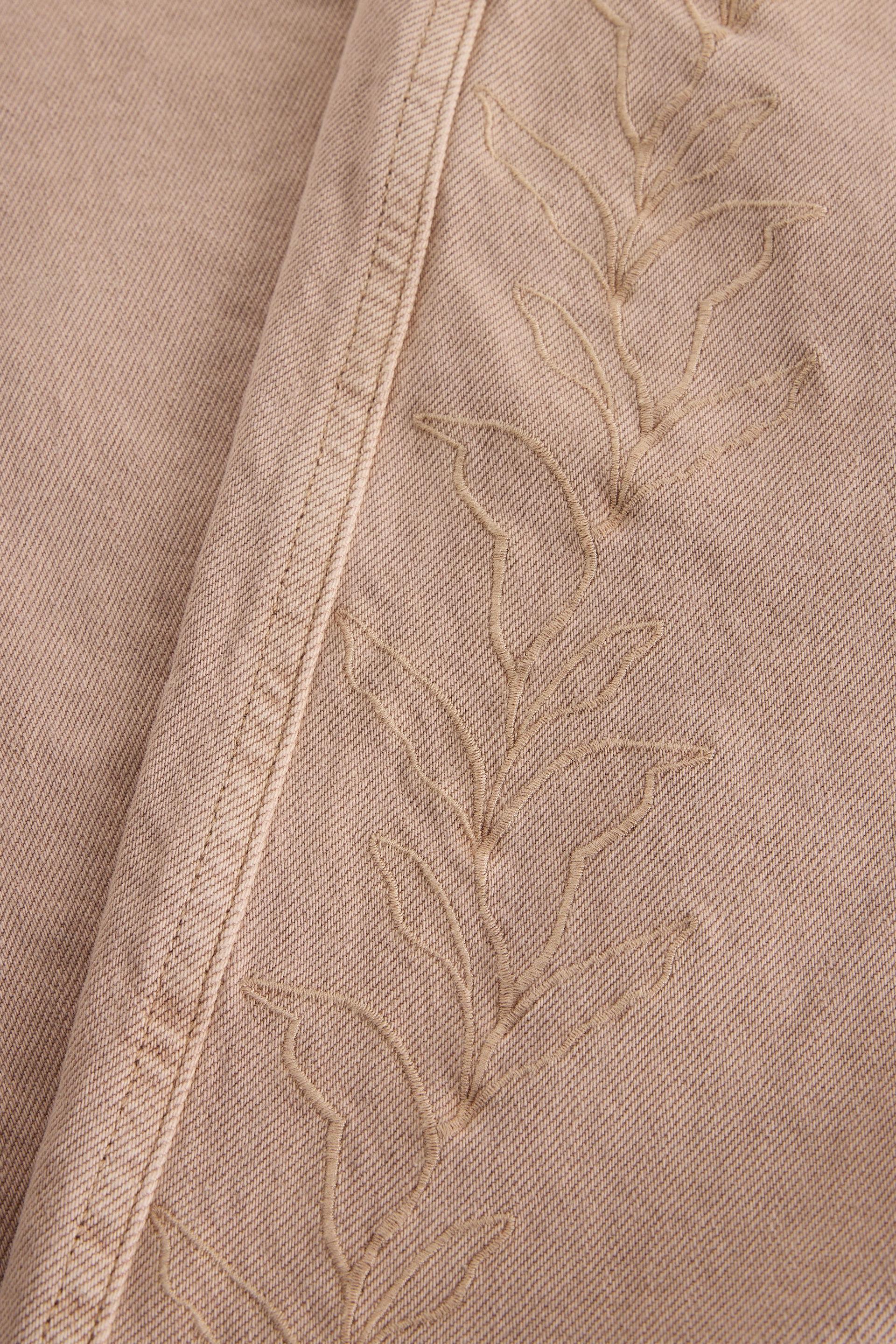 Close-up of light brown organic cotton denim fabric featuring delicate leaf embroidery and stitching details.