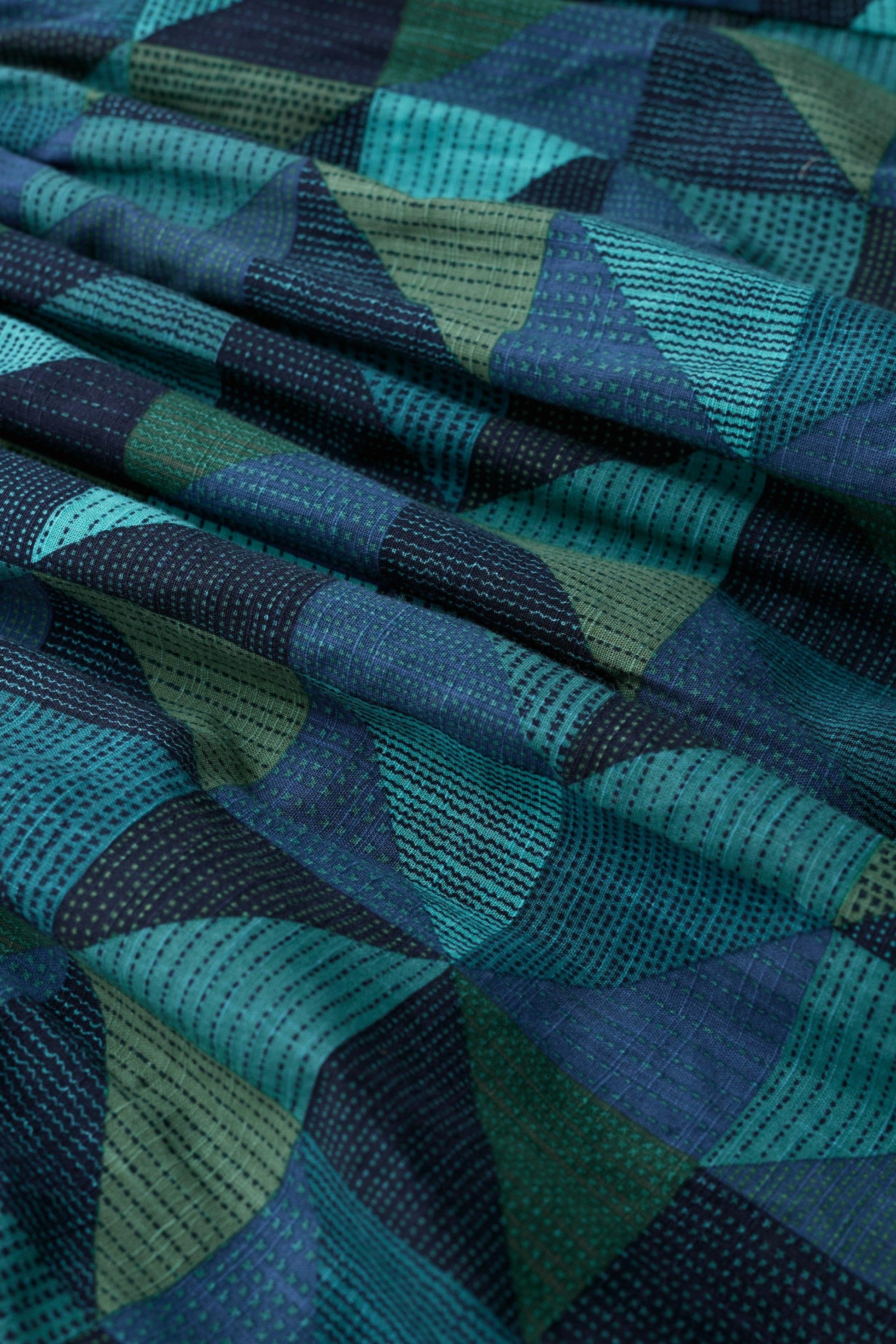 A close-up of draped fabric with a geometric pattern in shades of teal, blue, green, and dark navy, featuring dotted and textured woven sections.